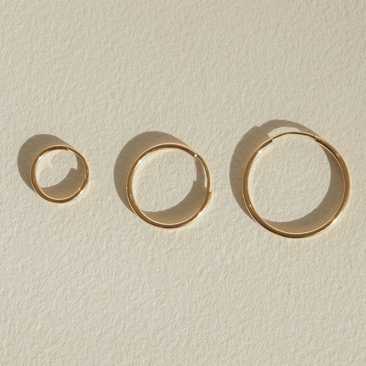 Four gold bangles of varying sizes displayed on a light textured surface, casting soft shadows, showcasing minimalist luxury jewellery design.