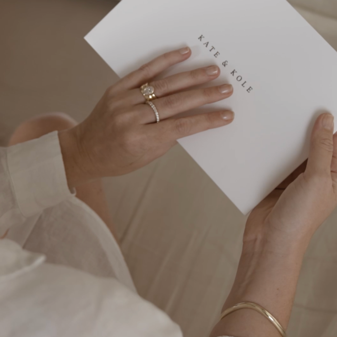 A person wearing elegant gold and diamond rings holds a KATE & KOLE branded envelope.