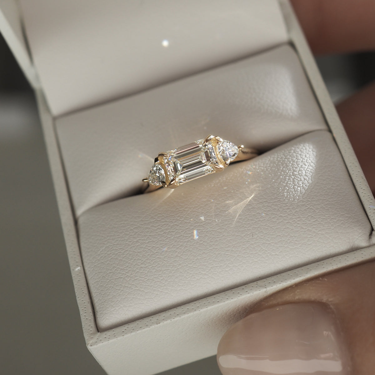 Emerald-cut diamond ring with two side stones in a beige velvet box, held by fingers; luxury jewellery, elegant design.