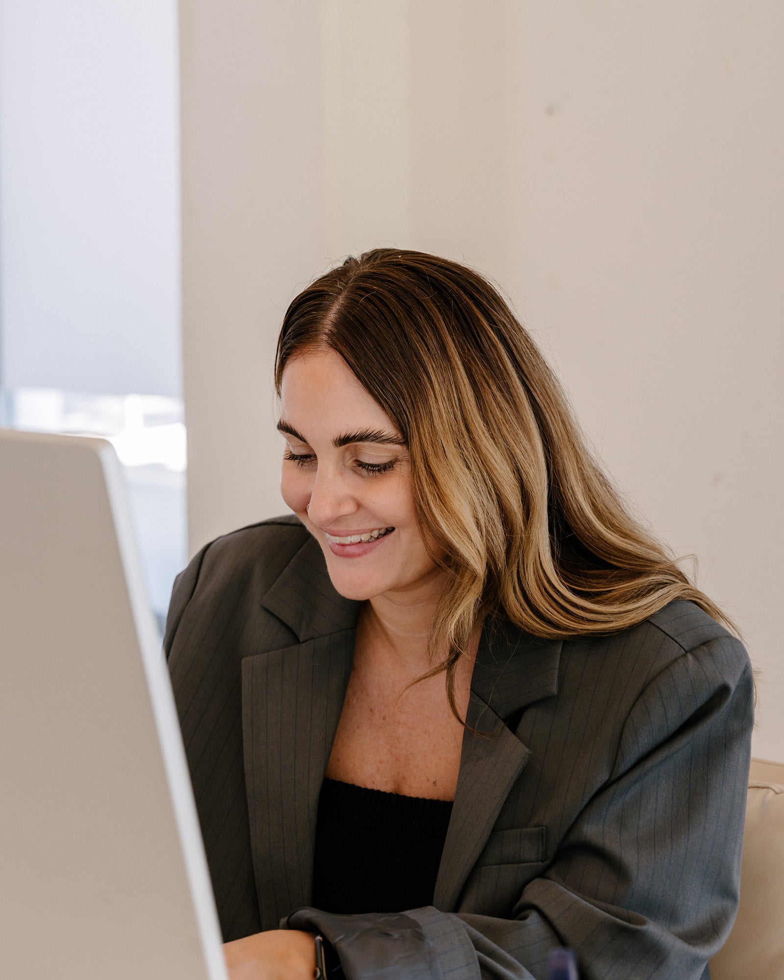 A woman in a grey blazer smiles while working on a laptop. She has long, straight hair and is seated indoors, focused on her screen.