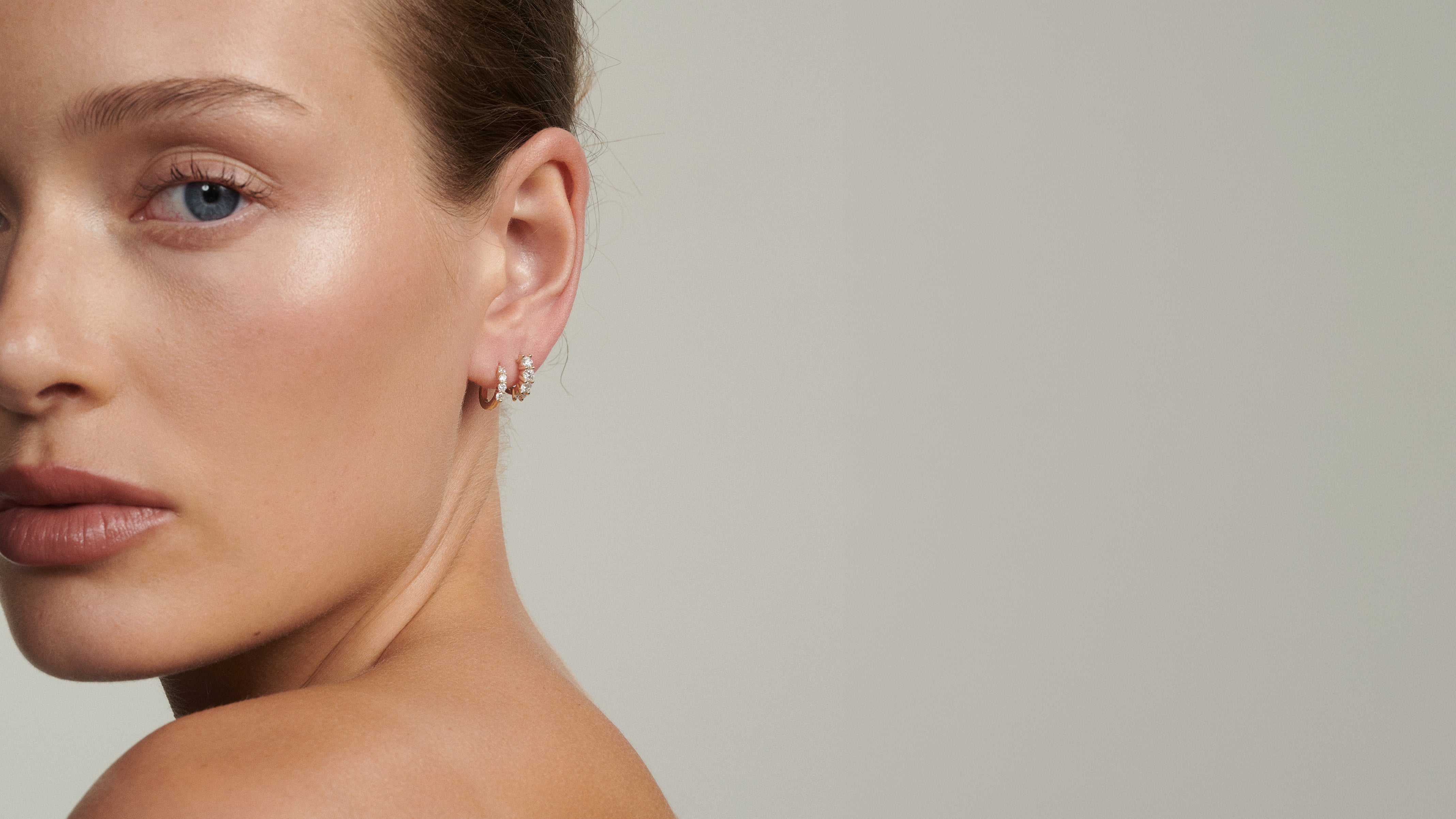Close-up of a woman's ear wearing two diamond huggies on a plain background