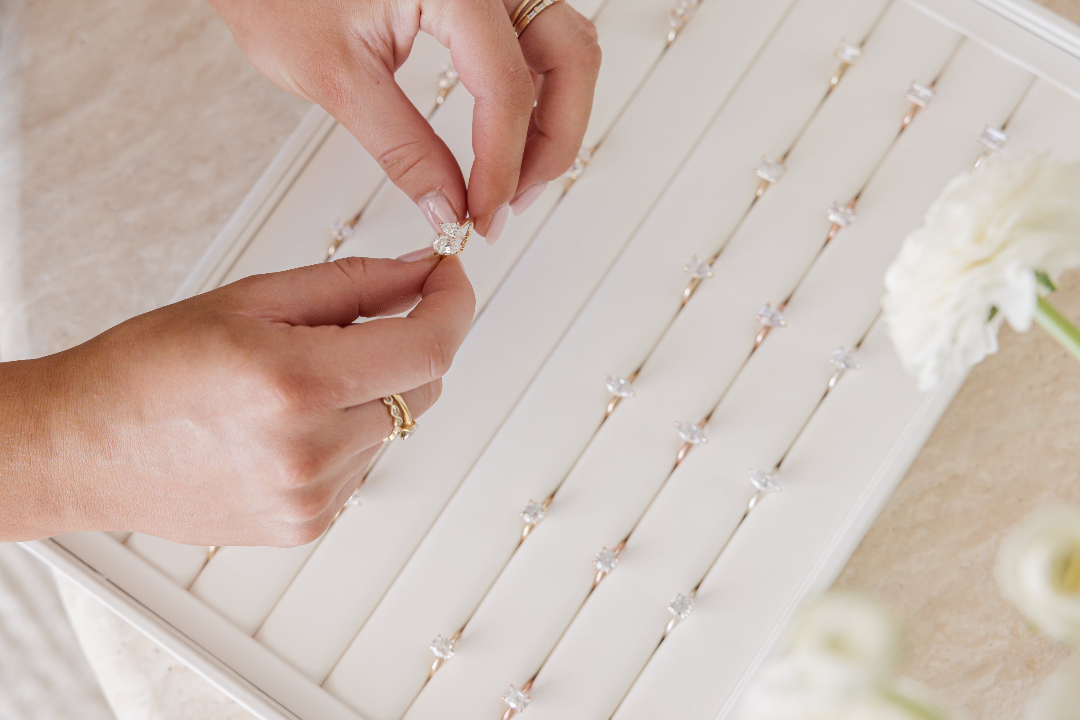 Hands selecting a diamond ring from a tray of elegant rings, displayed in a luxury jewellery setting with soft, neutral tones.
