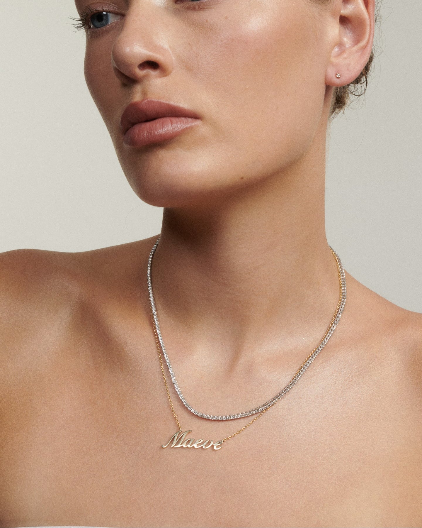 Woman wearing a gold necklace with 'Maeve' on a neutral background  [text overlay: Andrea wears our Name Plate Necklace & Tennis Necklace]