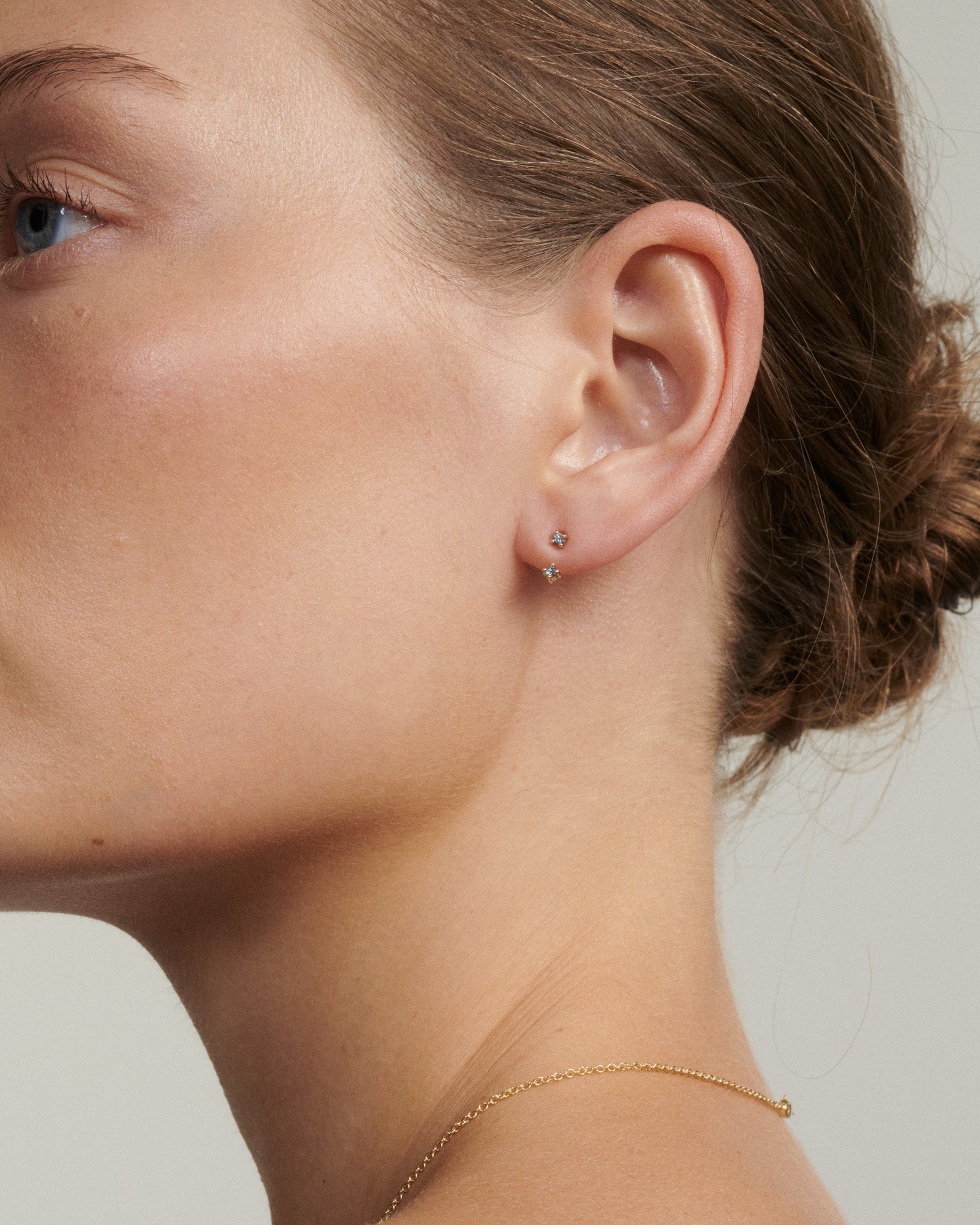 Close-up of a person wearing a small earring with a neutral background [text overlay: Andrea wears the Tiny Birthstone Jackets and Studs]