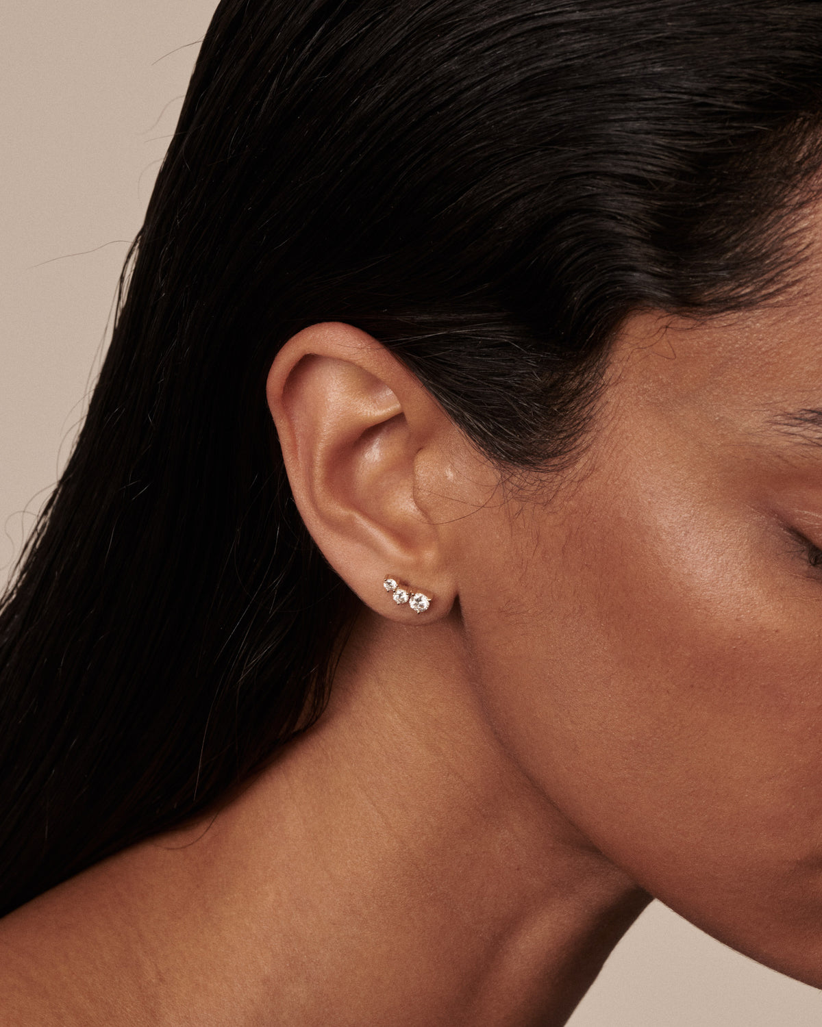 A close-up of a model's ear wearing a pair of small silver stud earrings with clear diamonds, set against a solid beige background.
