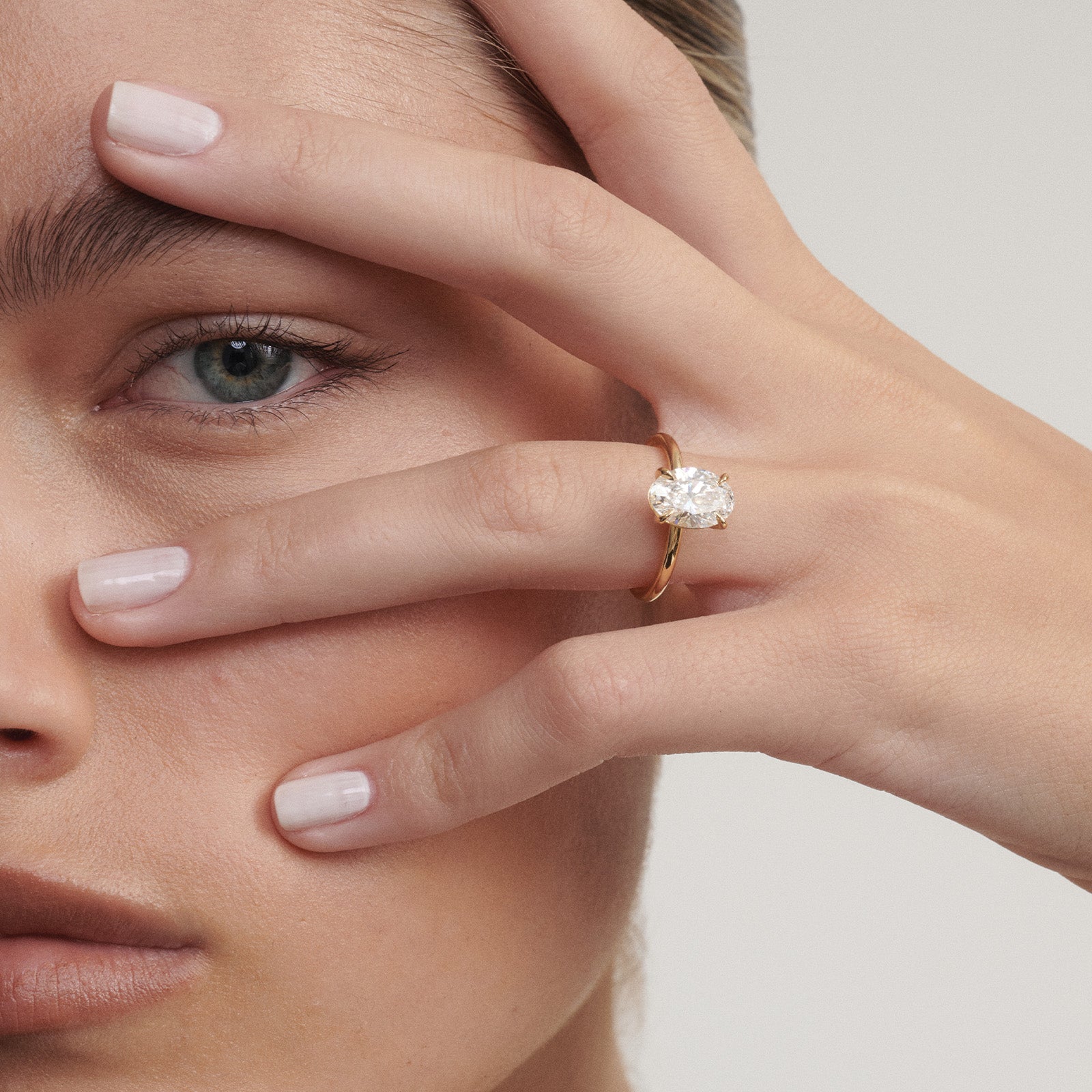 Close-up of a woman's face with an oval diamond ring on her finger against a neutral background