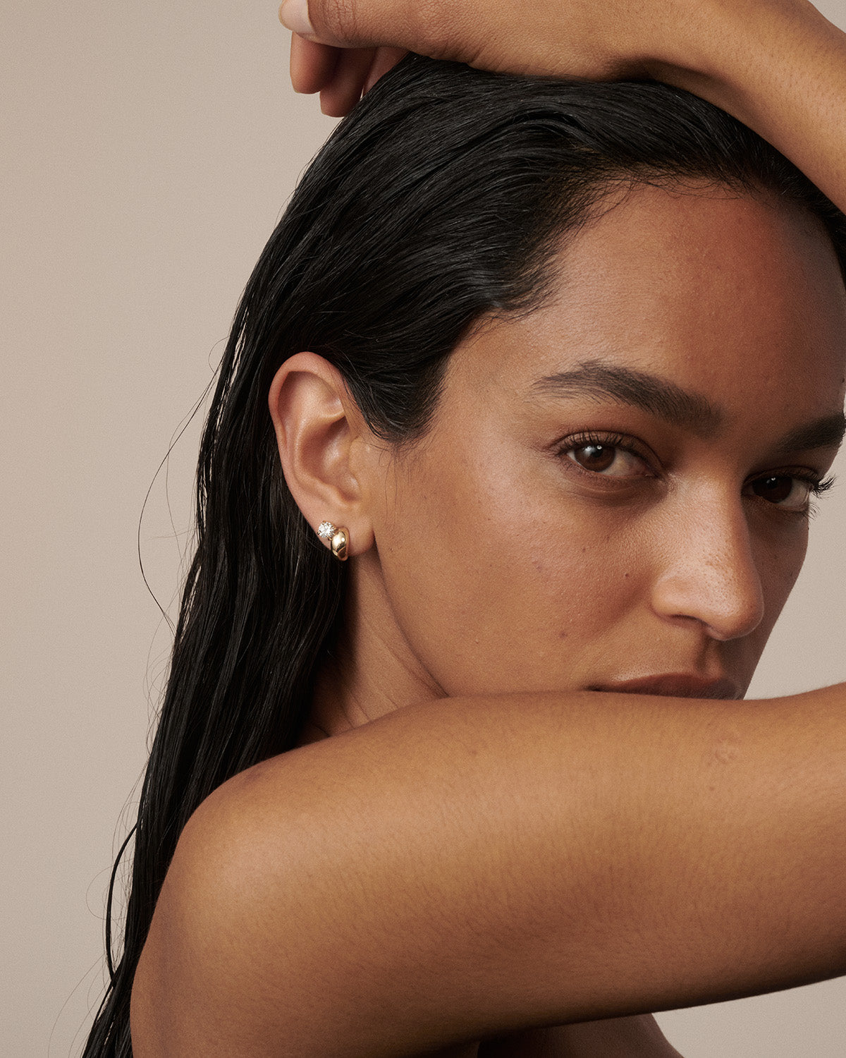 A close-up lifestyle shot of a woman with dark hair wearing a small gold stud earring, posing with her arm crossed over her face against a solid beige background. [text overlay: Francielle wears the bold style]