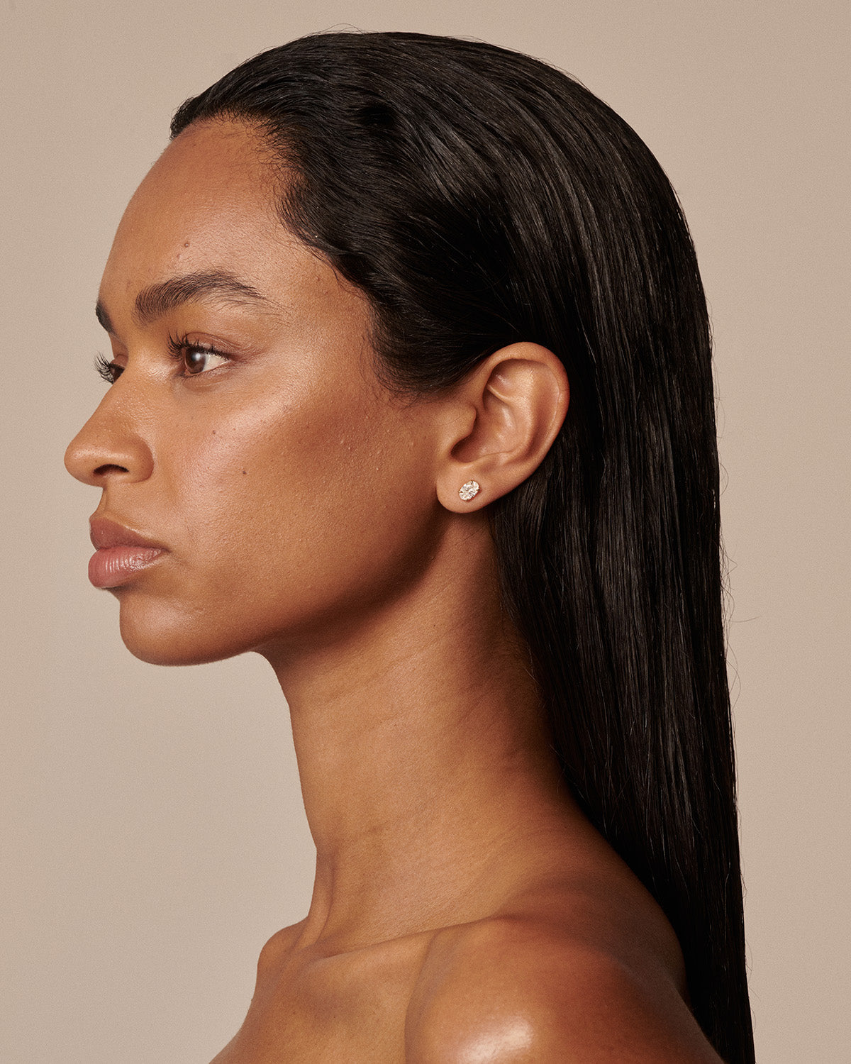 A close-up side profile of a woman with long, straight black hair wearing a small, round gold diamond stud earring against a solid beige background.