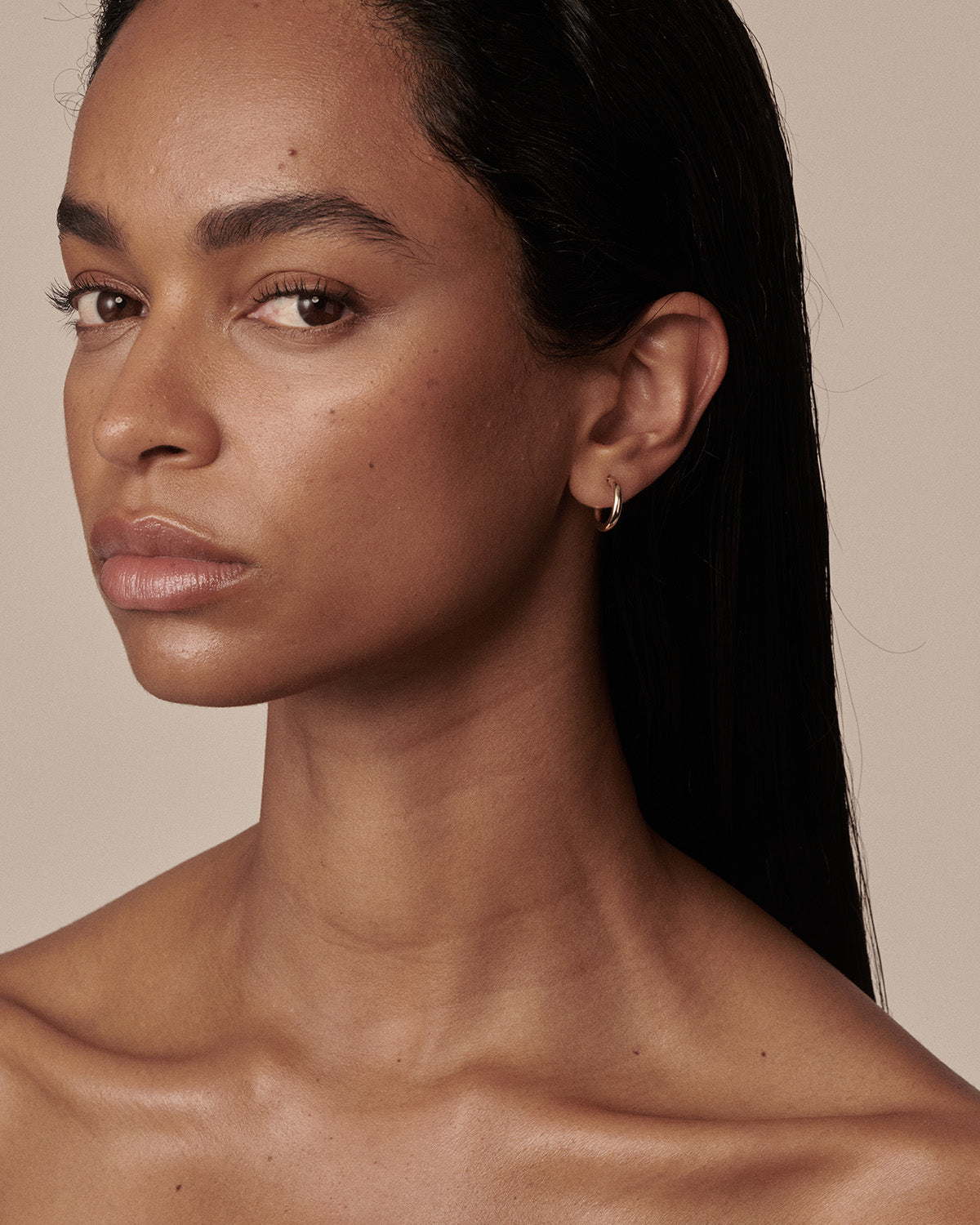 A close-up portrait of a woman with dark hair wearing a small gold hoop earring, set against a solid warm beige background.