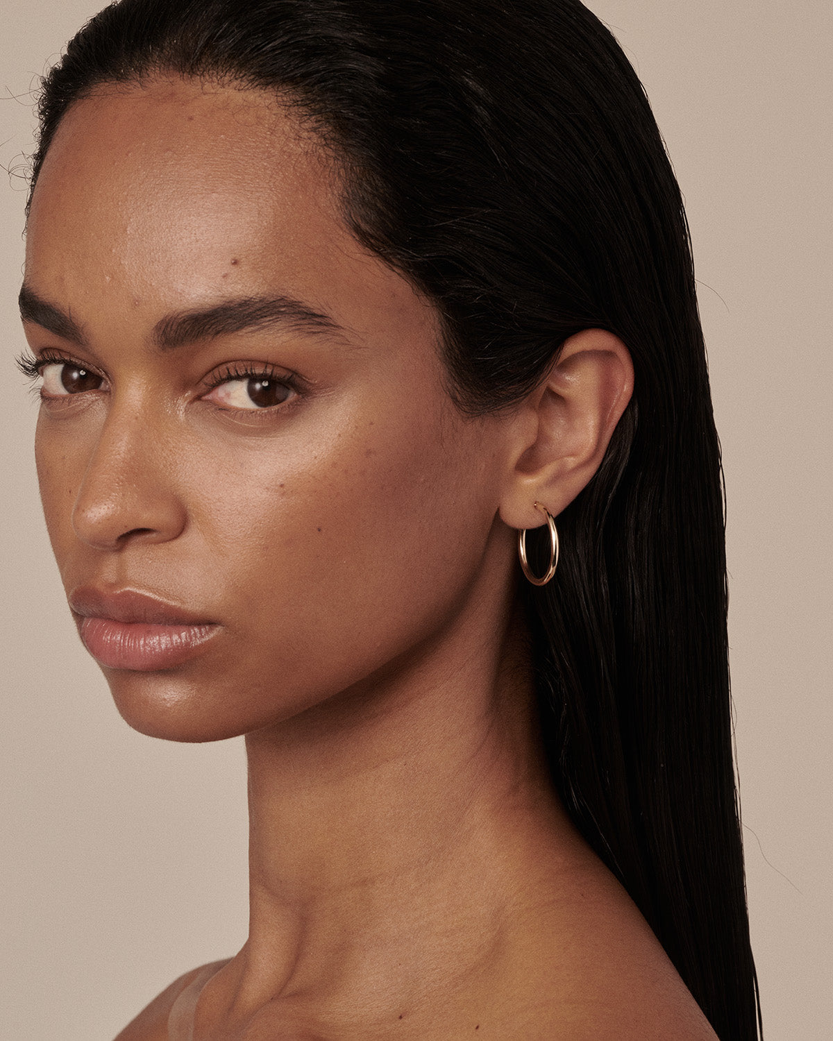 A close-up portrait of a woman with dark hair wearing a delicate gold hoop earring, set against a solid beige background.