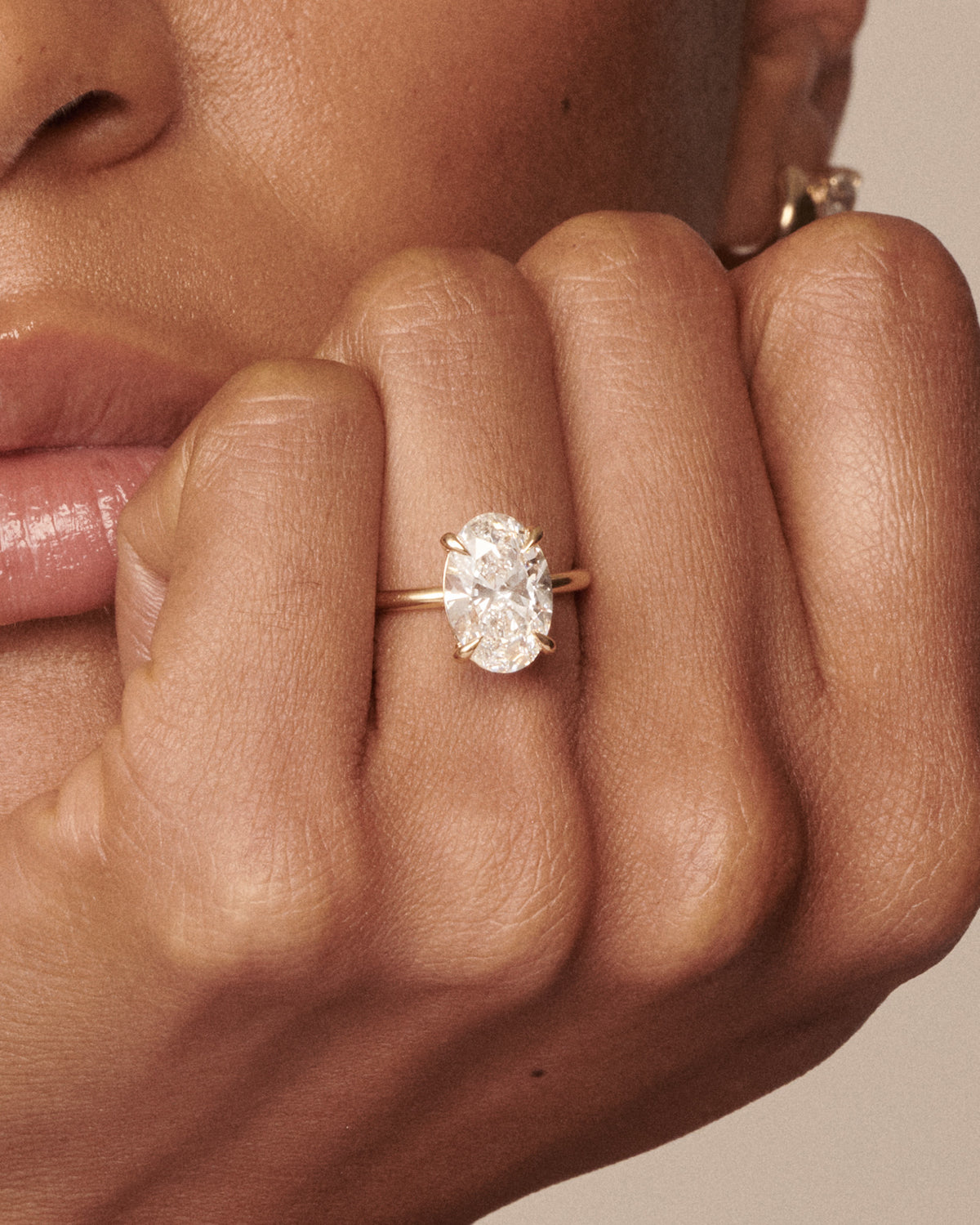 A close-up of a person's hand wearing a yellow gold ring with an oval-cut clear gemstone, set against a soft beige background.
