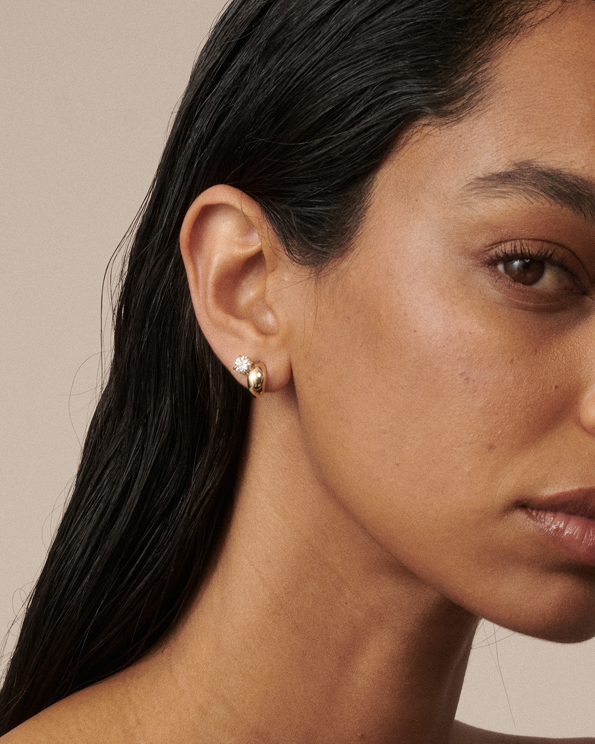 A close-up of a model's ear wearing a small gold huggie hoop earring with a tiny clear gemstone, against a neutral beige background.