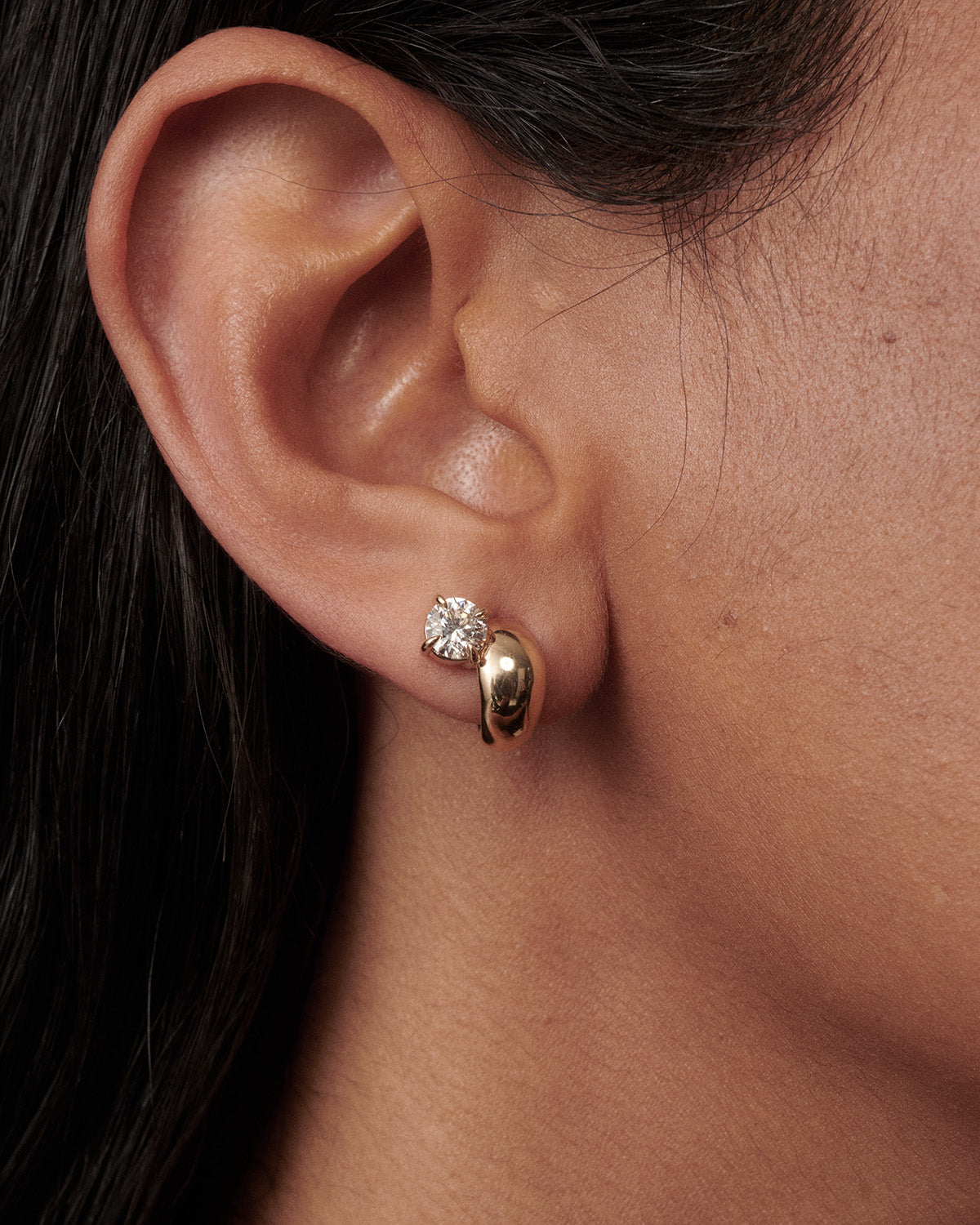 A close-up of a person's ear wearing a gold huggie hoop earring with a small clear gemstone stud above it.