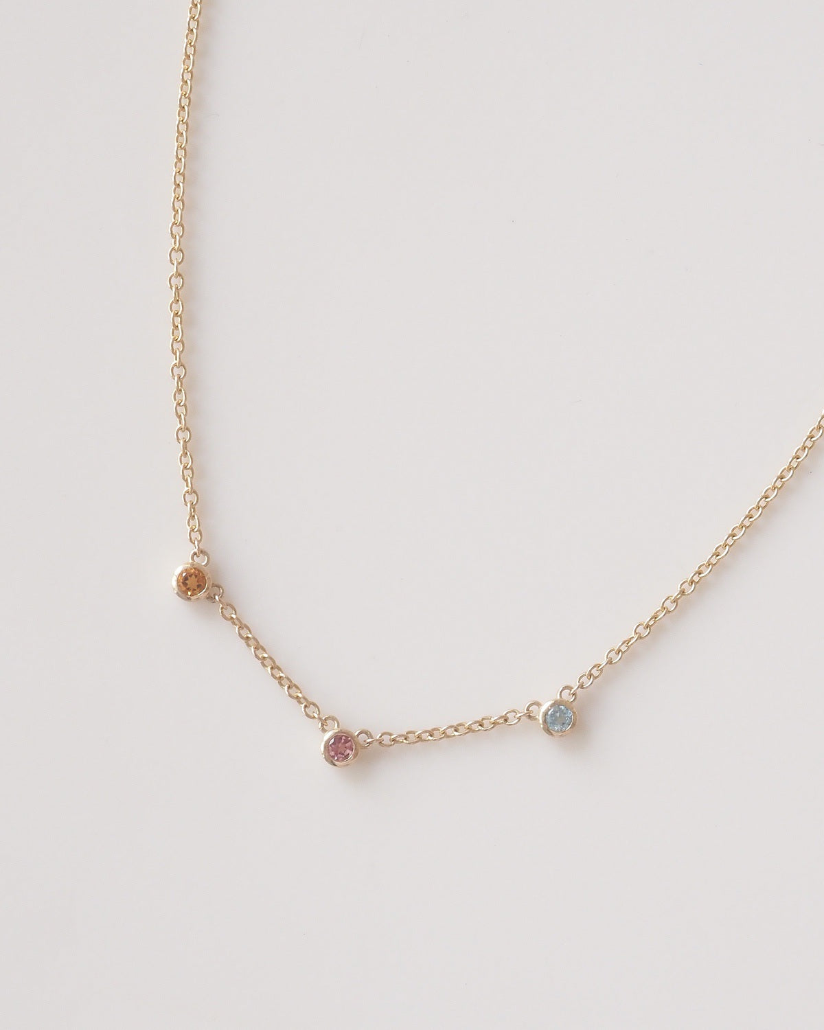 A delicate gold chain necklace featuring three small, spaced bezel-set gemstone charms in pink, blue, and orange, displayed on a clean off-white background.