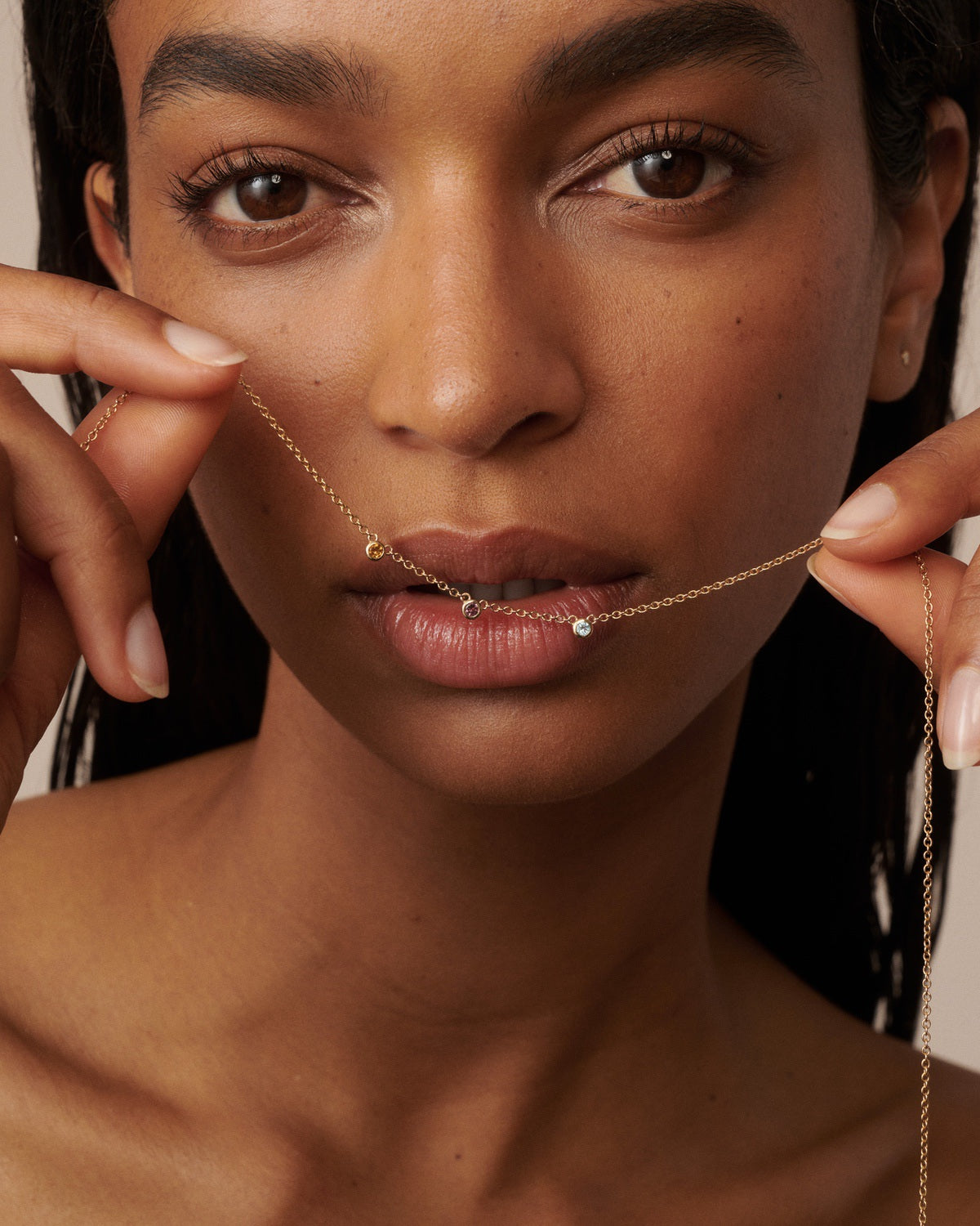 A delicate gold chain necklace with three small, individually colored gemstone beads (citrine, pink tourmaline, and diamond) worn on a model's neck held across the models lips.