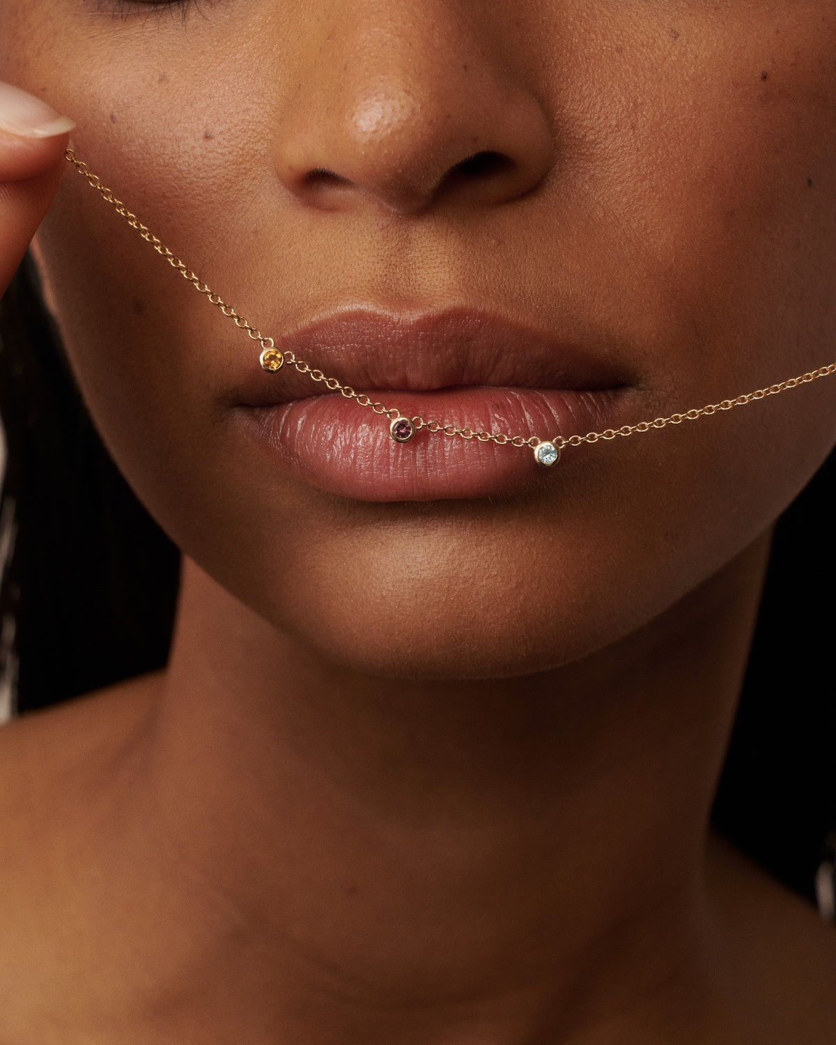 A close-up of a model wearing a delicate gold chain necklace draped across her lips, featuring small gold and clear gemstone beads.