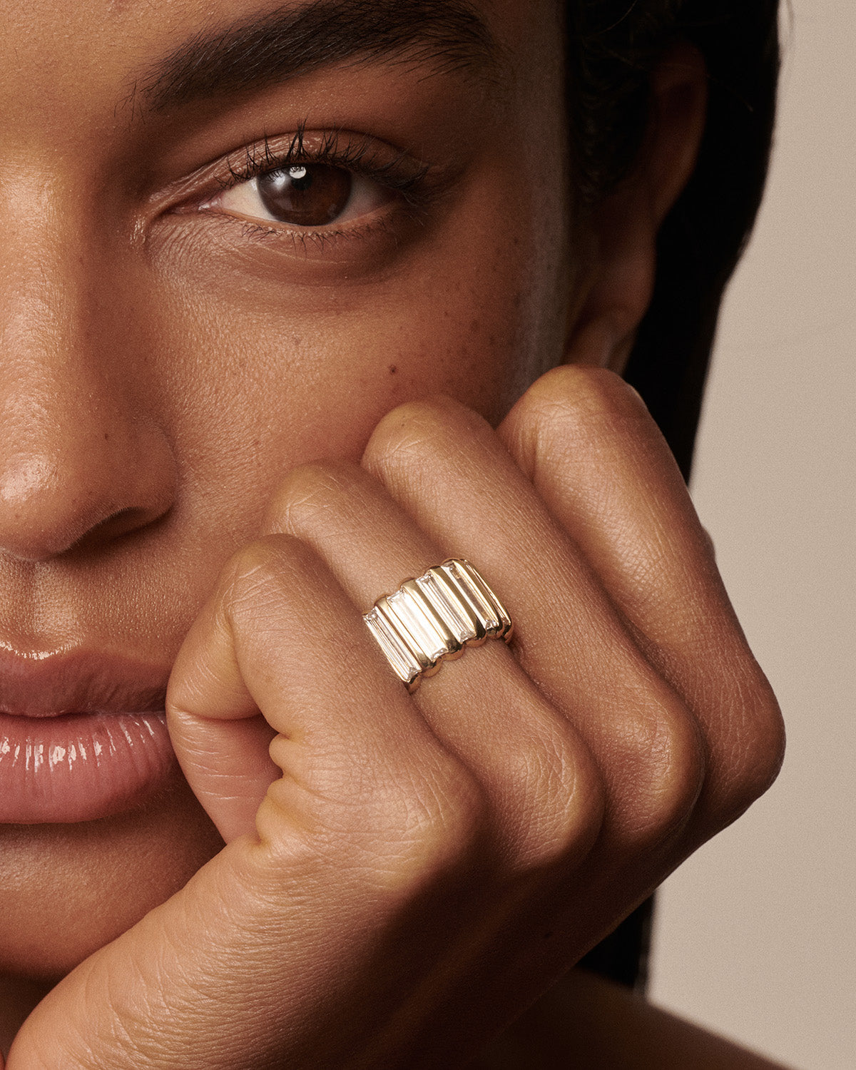 A close-up lifestyle shot of a model wearing a wide, ribbed gold ring on her finger, with her face and hand prominently featured against a neutral beige background.