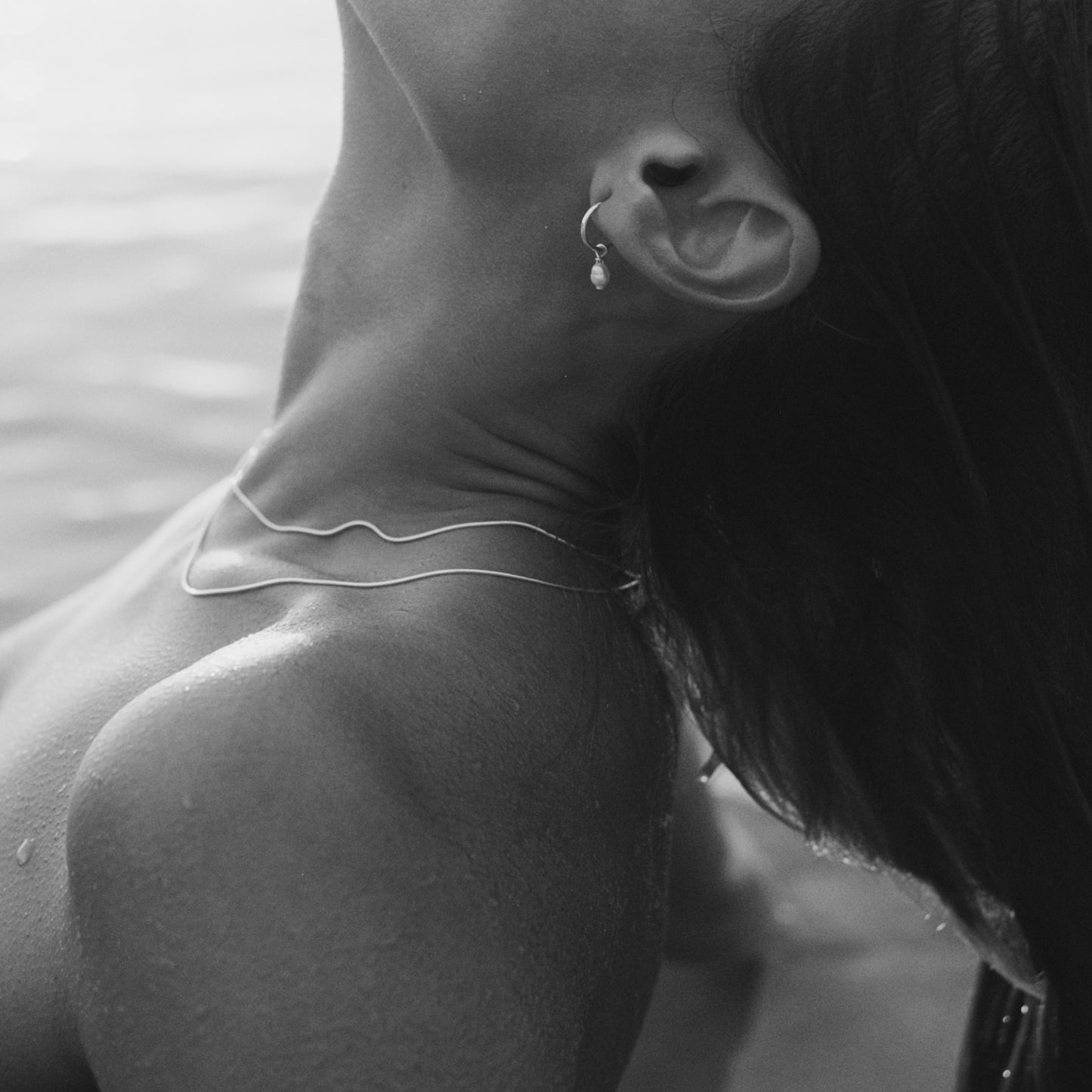 Monochrome image of a woman's neck adorned with a delicate necklace and earring, water droplets visible, evoking luxury and elegance.