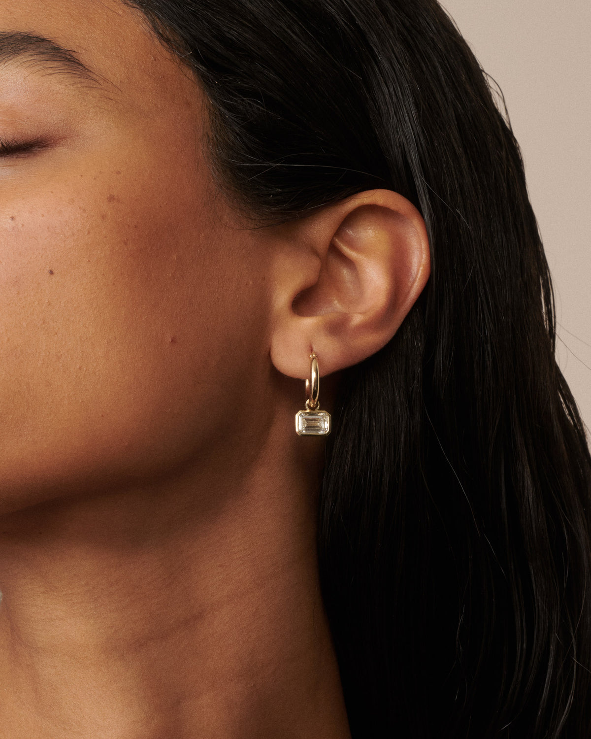 A close-up of a model's ear wearing a delicate gold huggie hoop earring with a small clear faceted rectangular drop stone, set against a warm beige background. 
