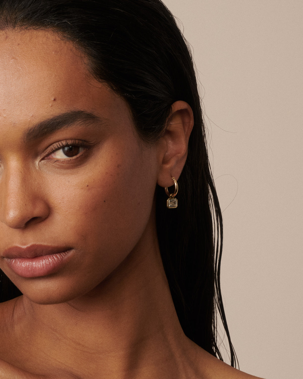 A close-up portrait of a woman with dark hair wearing small gold hoop earrings with clear diamond drops, set against a solid beige background. 