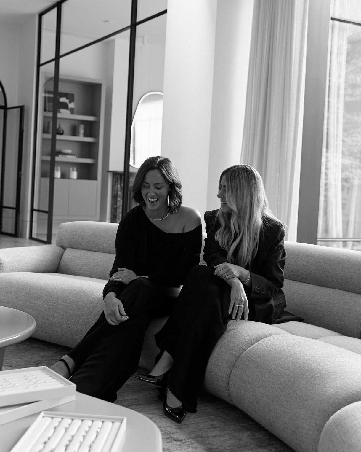 Two women in elegant attire sit on a plush sofa in a modern, luxurious living room, engaged in conversation, exuding style and sophistication.