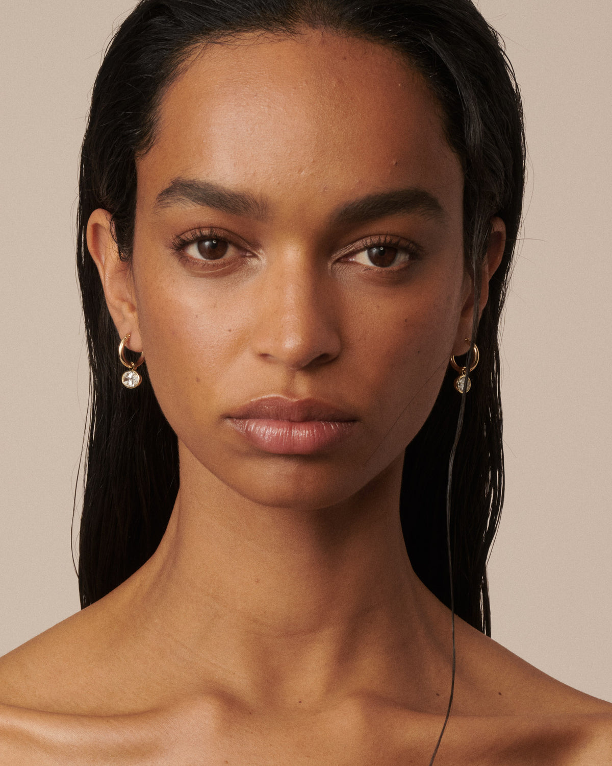 A close-up portrait of a woman with dark hair wearing small gold hoop earrings with clear diamond drops, set against a solid beige background. 