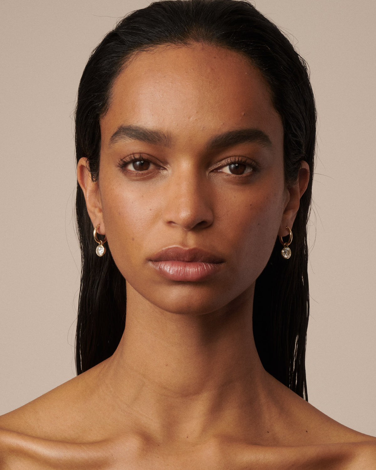 A close-up portrait of a woman with dark hair wearing small gold hoop earrings with clear diamond drops, set against a solid beige background. 
