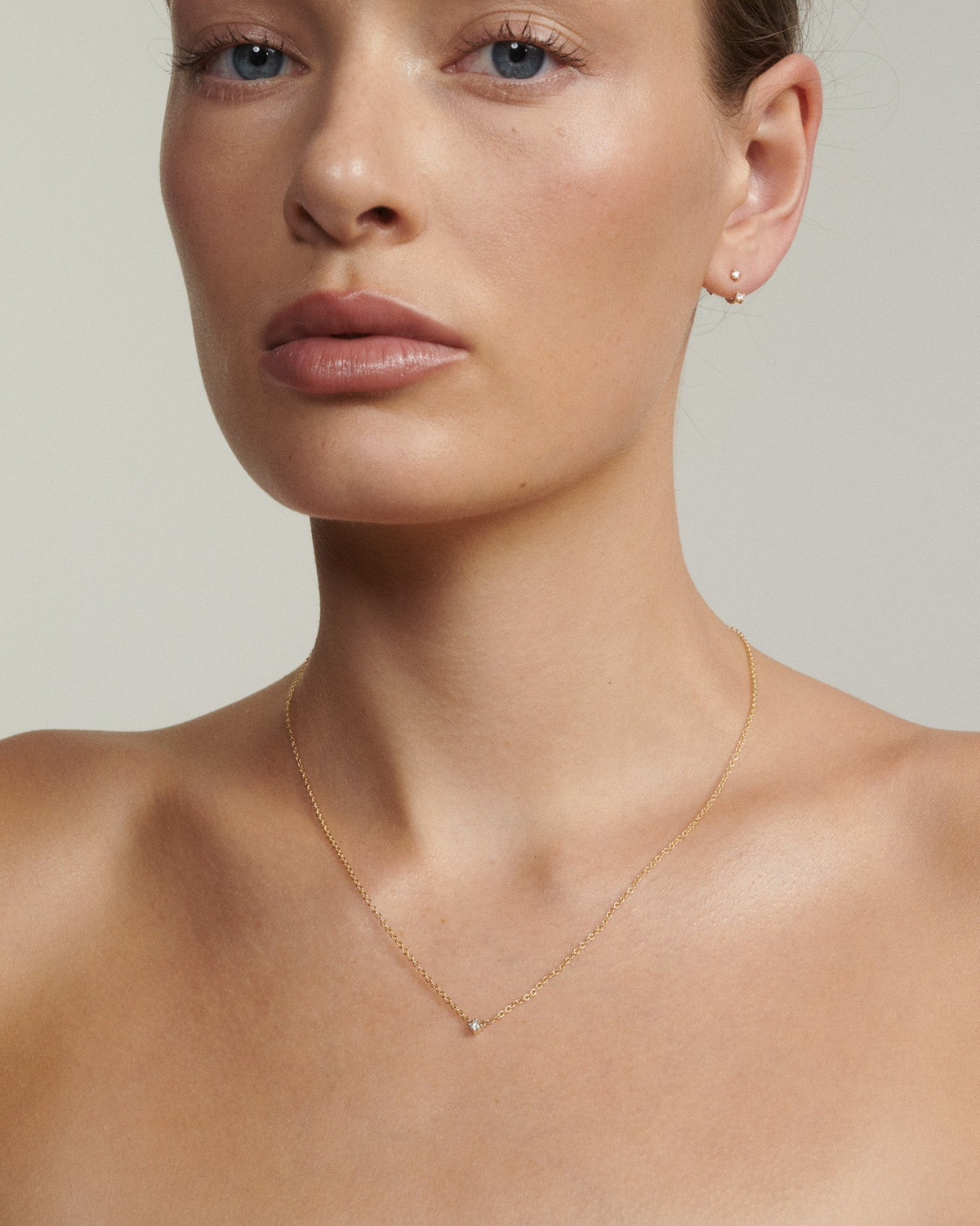 Close-up of a person wearing a delicate gold necklace against a neutral background [text overlay: Andrea wears the Tiny Birthstone Necklace, Studs & Jackets]