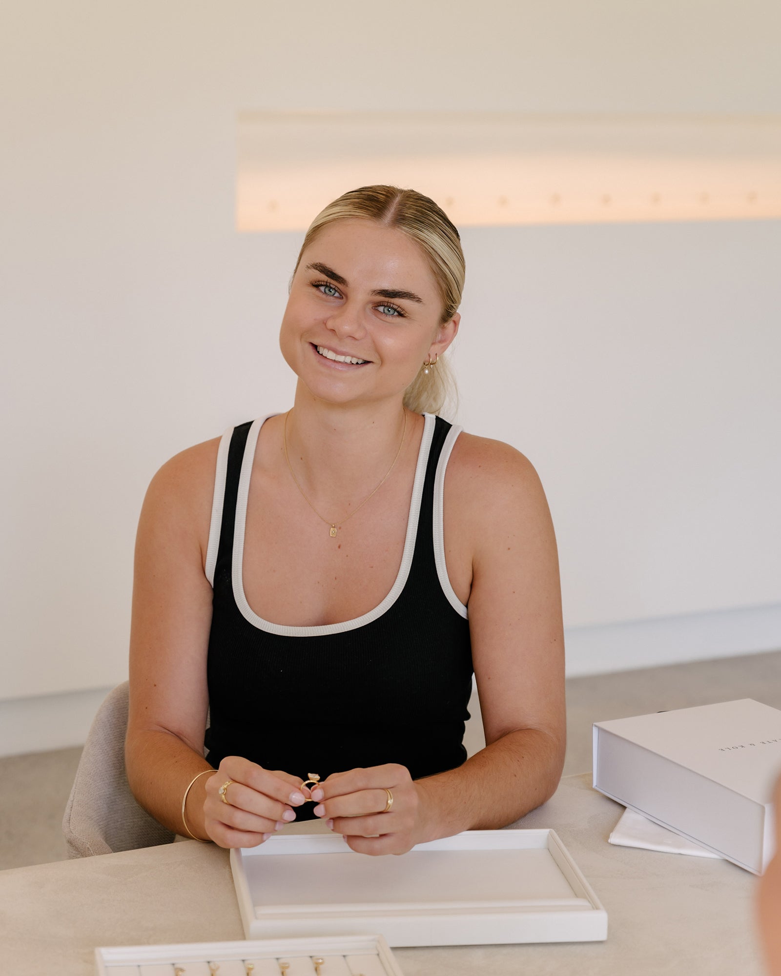 Smiling woman seated at a table, handling jewellery pieces in a well-lit room. She wears a black top, accessorised with simple, elegant jewellery.