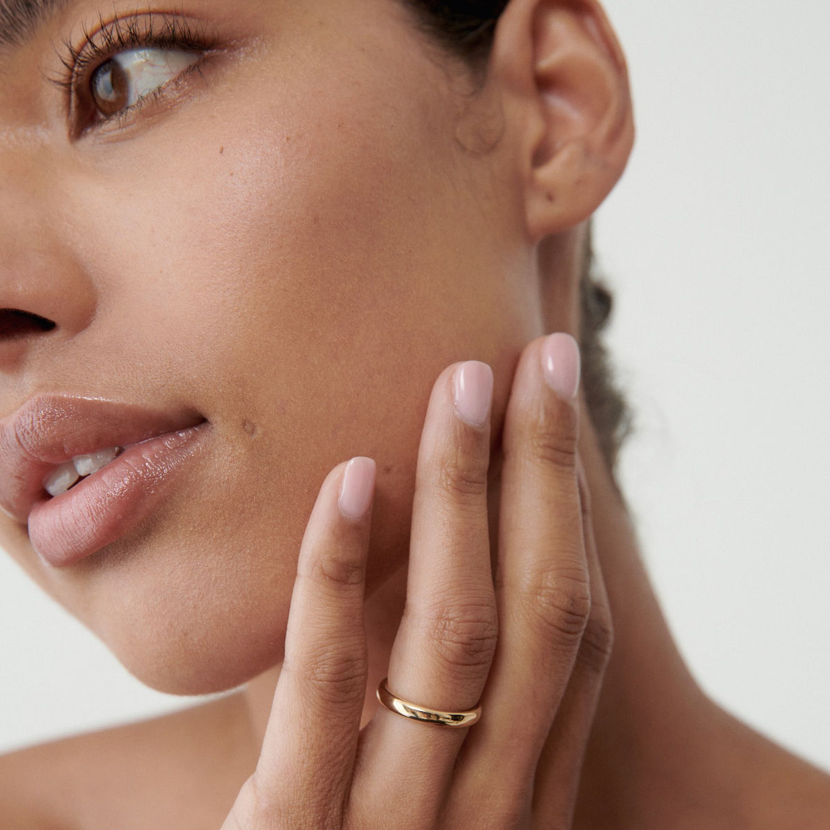 Close-up of a woman's face and hand, showcasing a minimalist gold ring on her finger, with a soft pink nail polish. Luxe jewellery detail.