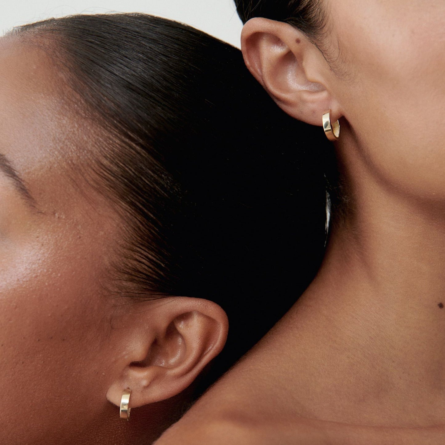 Two women wearing elegant gold hoop earrings, showcasing minimalist luxury jewellery against smooth skin and a neutral background.