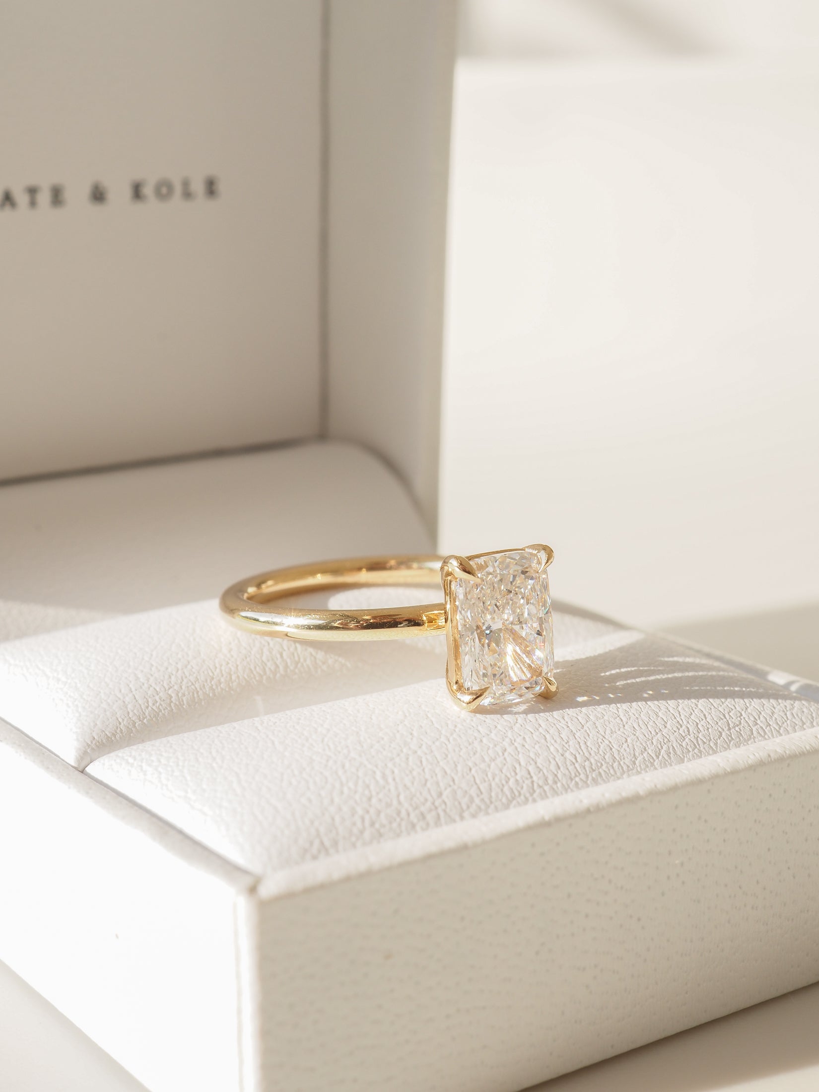 Elegant gold ring with a radiant-cut diamond displayed on a white leather cushion box. Perfect engagement jewellery, luxurious and sophisticated.