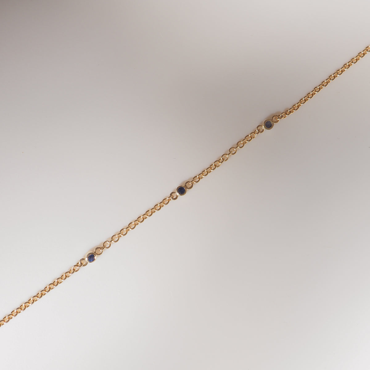 Gold chain bracelet with round links and scattered blue gemstones on a light background. Luxurious jewellery design.