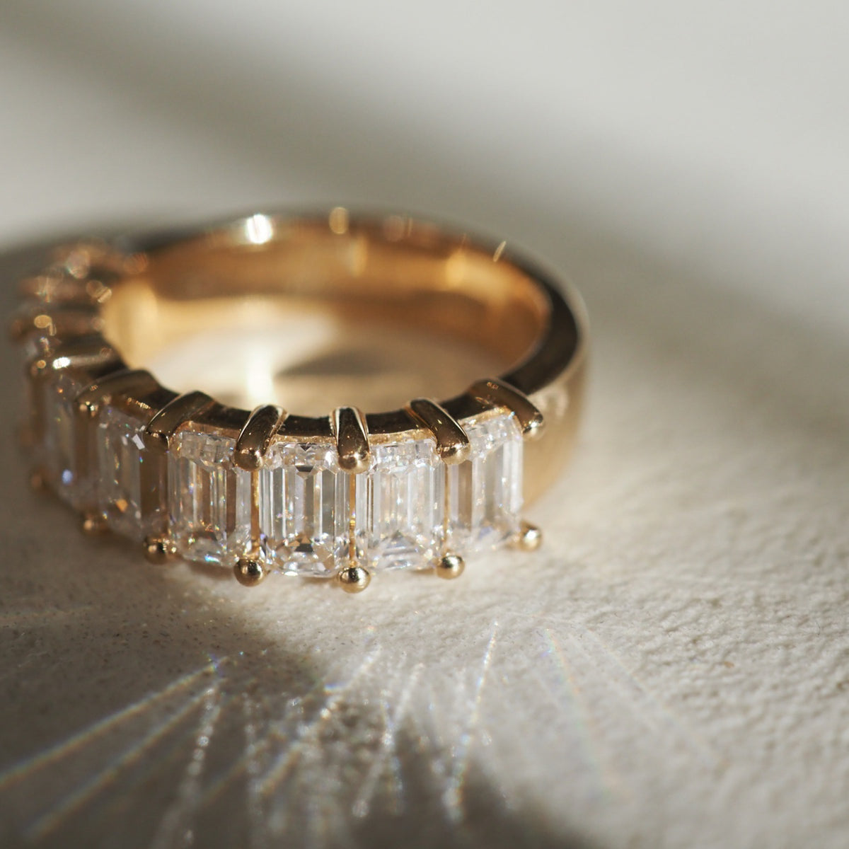 Gold ring with rectangular cut diamonds on a textured surface, catching light reflections. Luxury jewellery in an elegant setting.[text overlay: Emerald Ring]