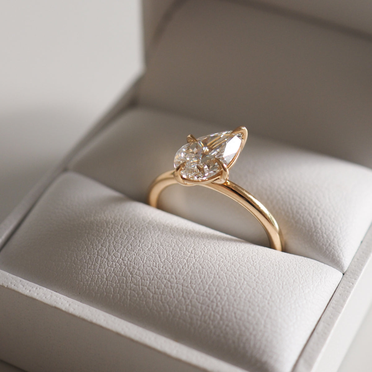 Gold solitaire engagement ring with pear-shaped diamond set on a plush white cushion in an elegant box. Luxurious jewellery design.
