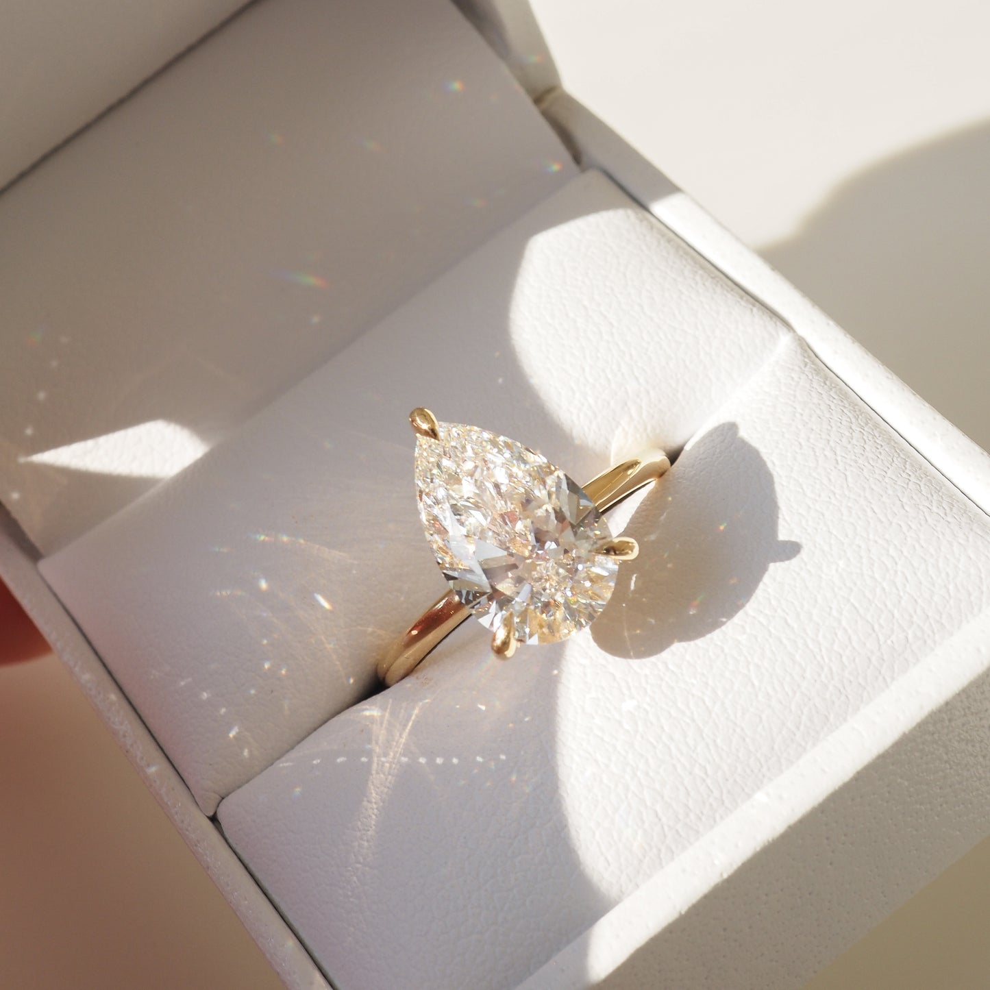 Pear-cut diamond ring in gold setting, displayed in a white jewellery box. Sunlight creates rainbow reflections, emphasising luxury and elegance.