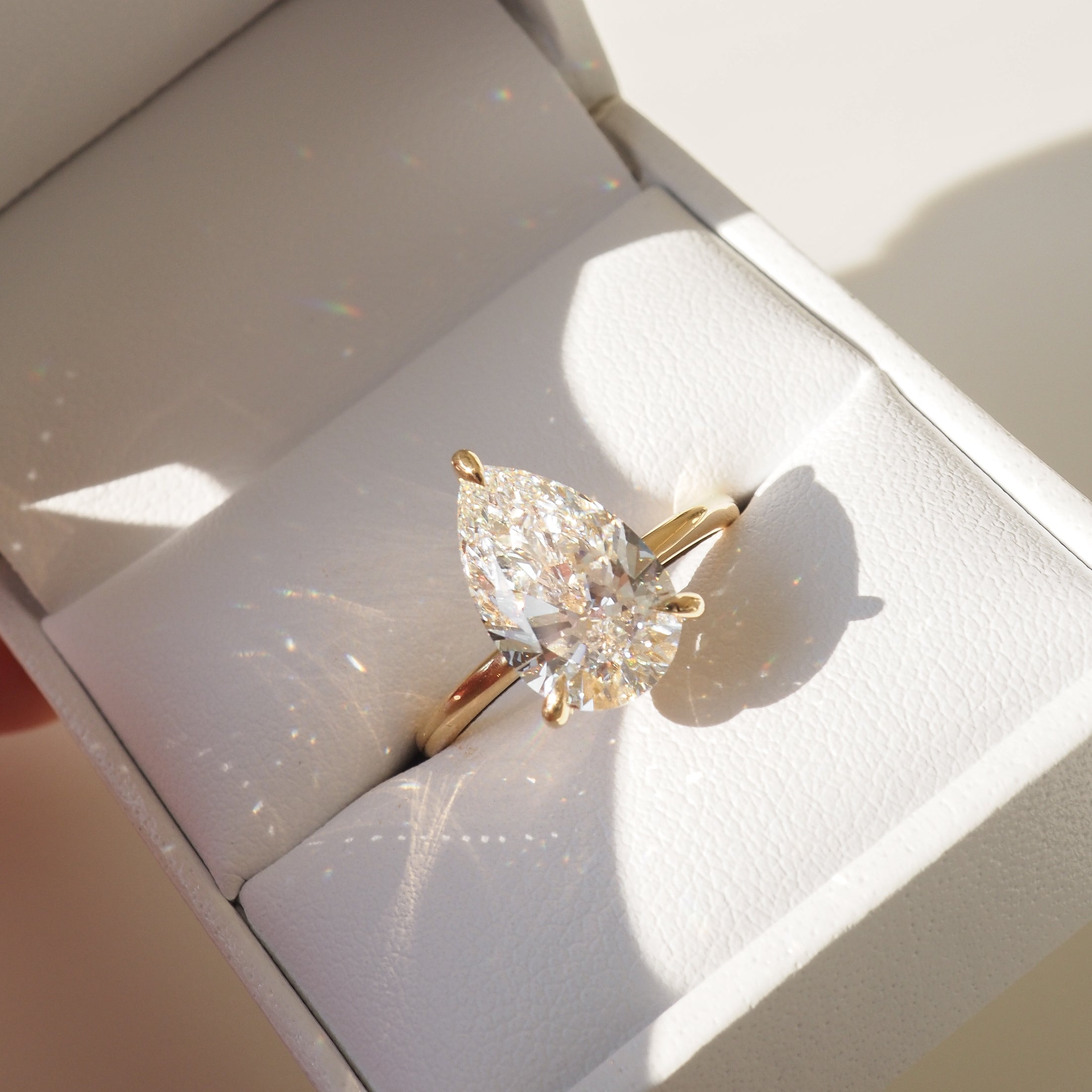 Pear-cut diamond ring in gold setting, displayed in a white jewellery box. Sunlight creates rainbow reflections, emphasising luxury and elegance.