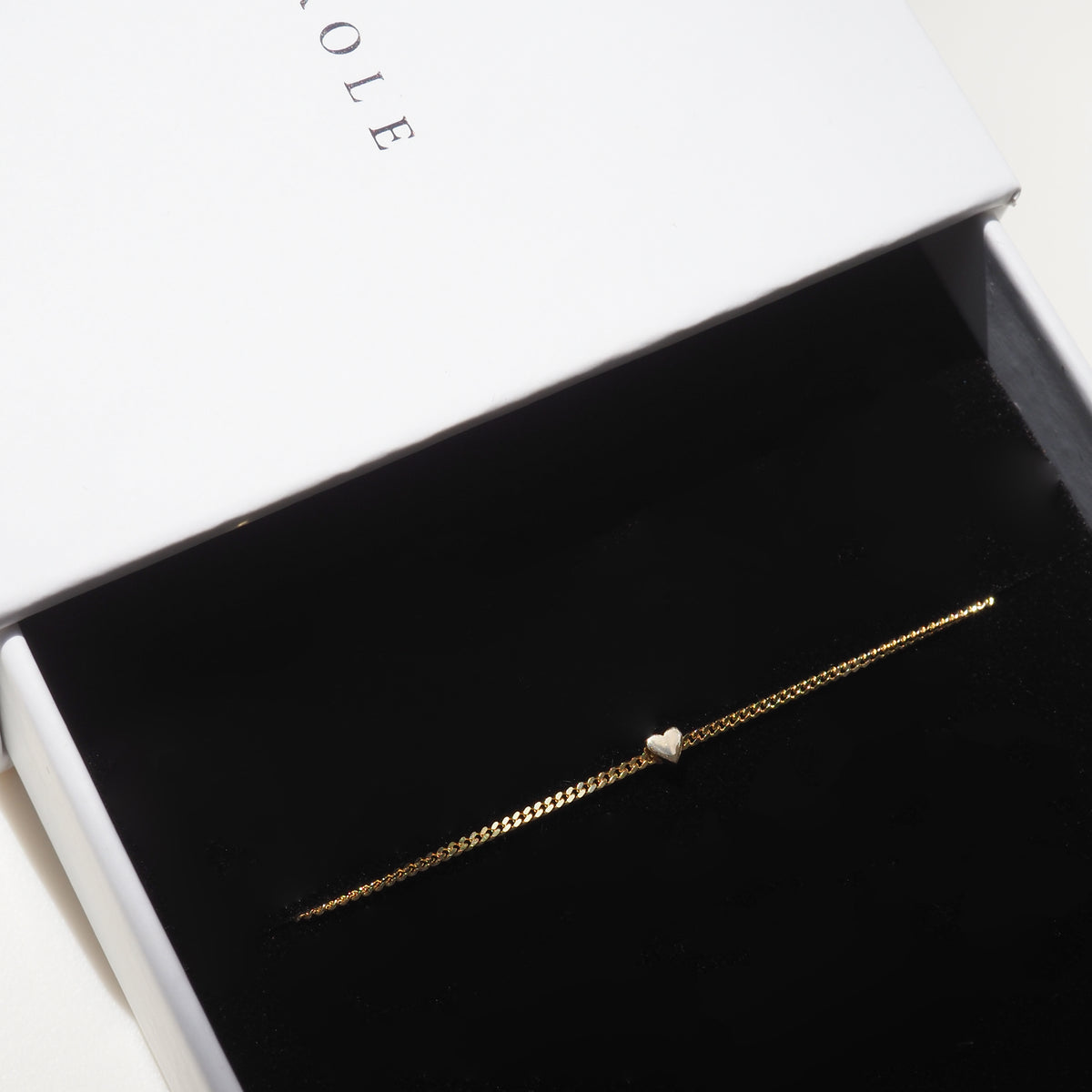 Gold bracelet with a heart charm in a black velvet box, partially open white box lid above. Luxury jewellery packaging.