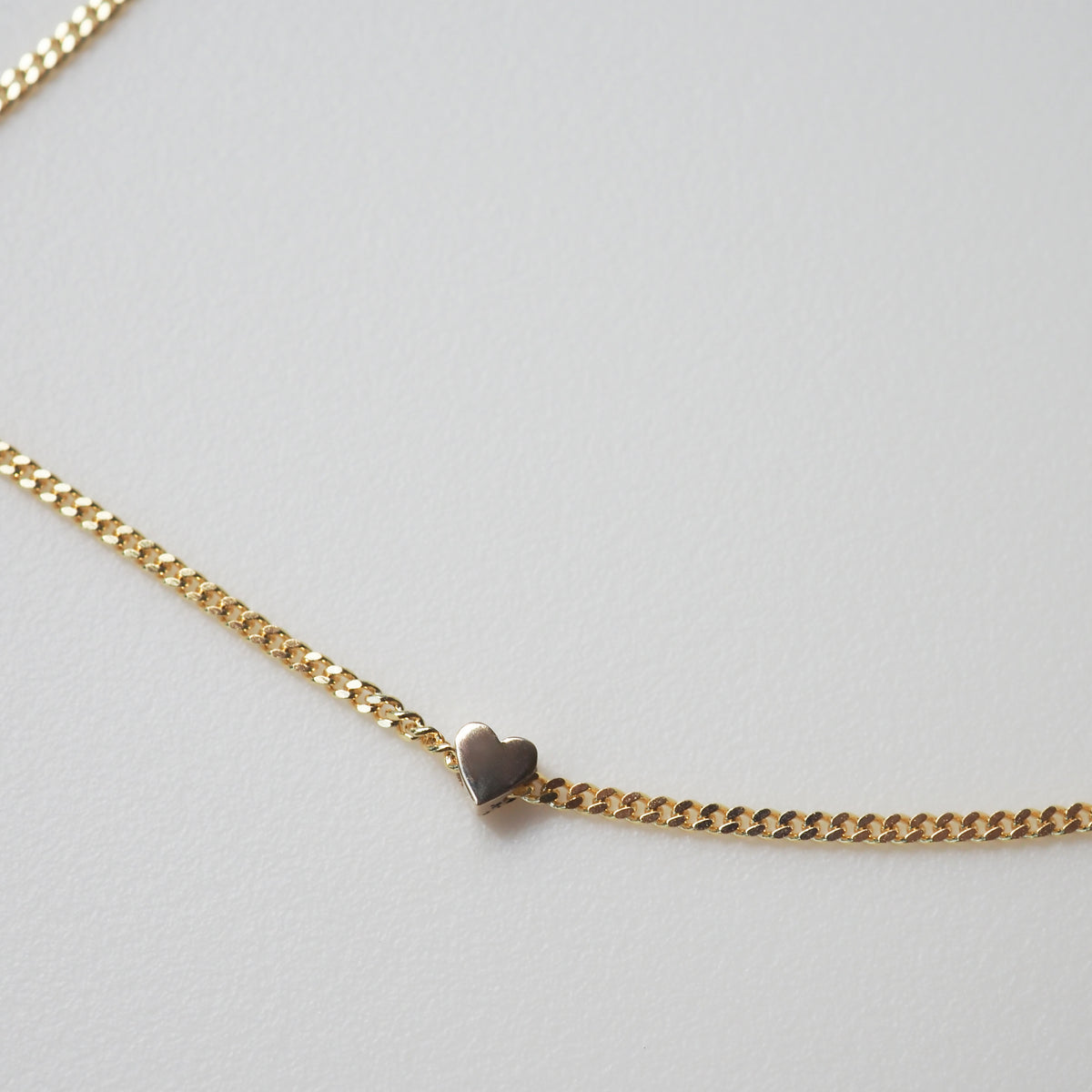 Gold chain bracelet with a small heart charm on a light grey background. Ideal for luxury jewellery lovers.