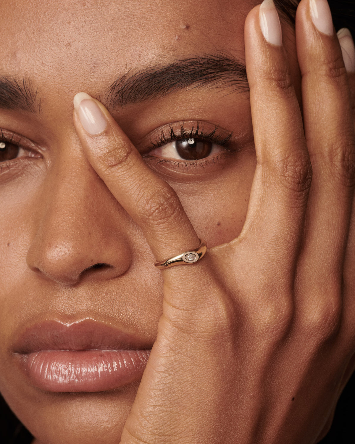 A close-up lifestyle shot of a model's face and hand, wearing a delicate gold ring with a small oval clear diamond on the ring finger.