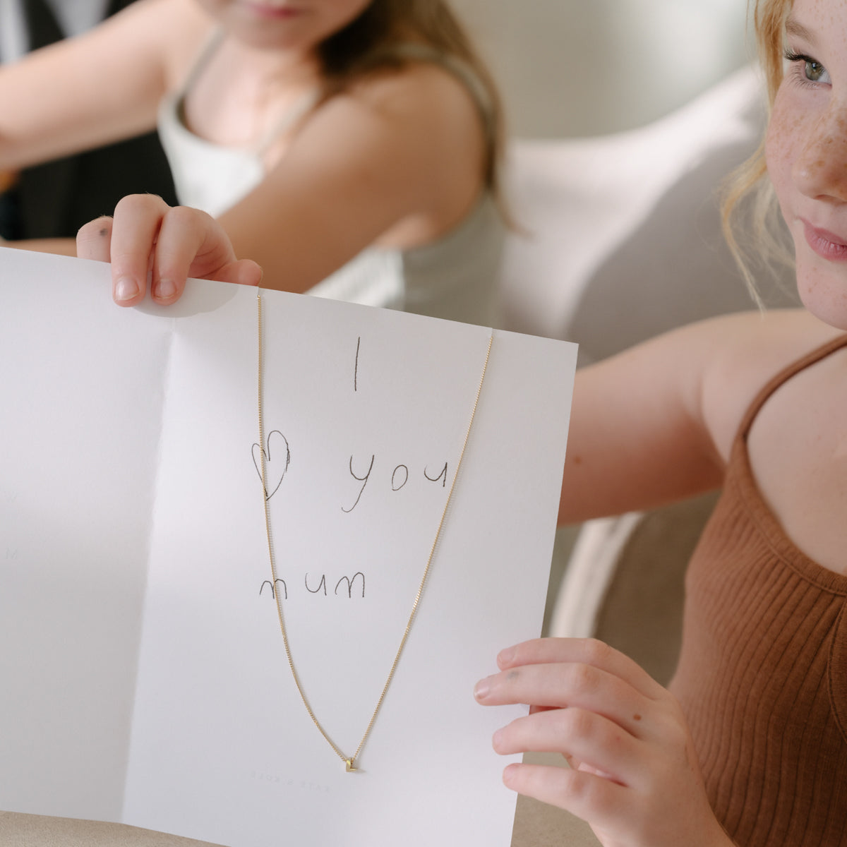 Child holding card with handwritten "I love you mum" and delicate gold necklace, perfect for Mother's Day or luxury jewellery gift.