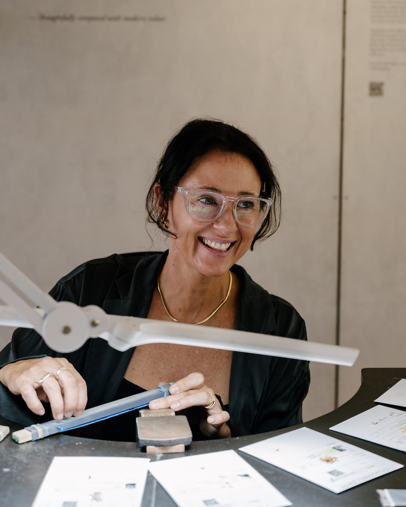 Smiling jeweller working at a bench, wearing glasses, holding a tool. Jewellery sketches and tools visible. High-end craftsmanship setting.