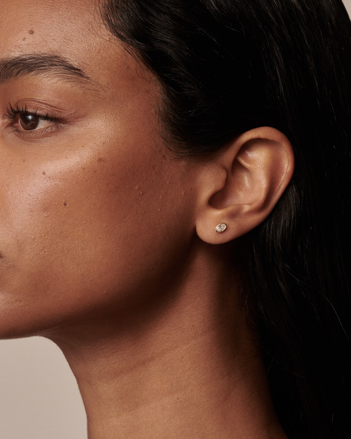 Close-up of a model's ear wearing a small, round, bezel-set clear gemstone stud earring.
