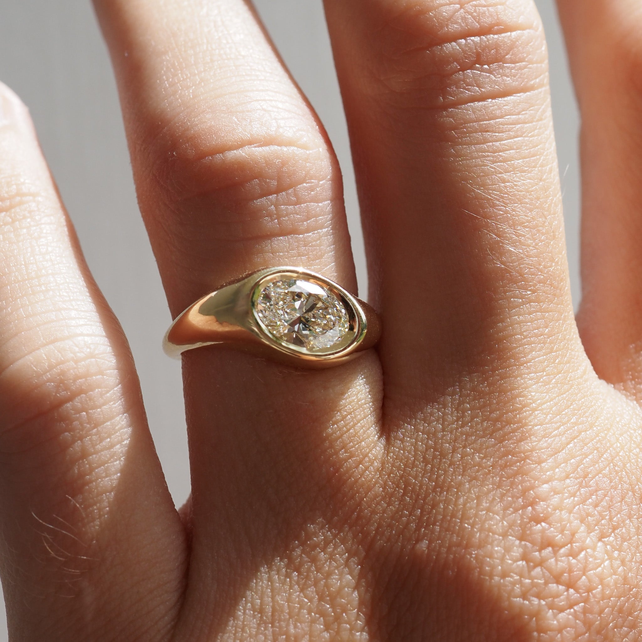 Gold ring with a diamond on a person's finger against a neutral background [text overlay: 1.5ct Riva Signet]