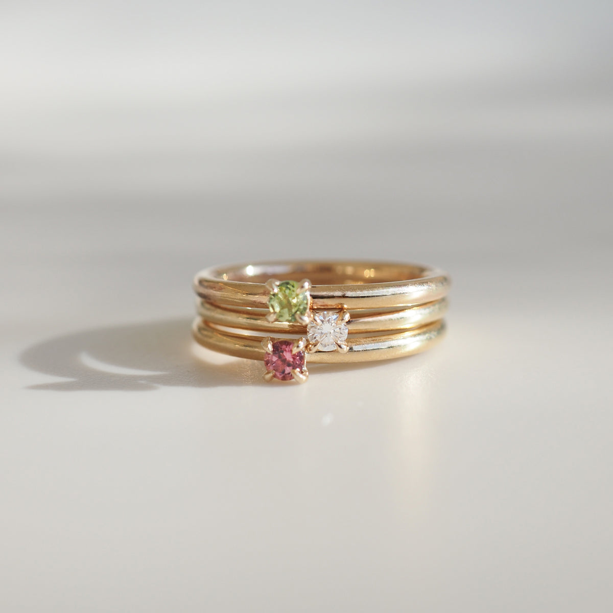 Three gold stacking rings with green, pink, and clear gemstones elegantly displayed on a light surface. Luxury jewellery design.