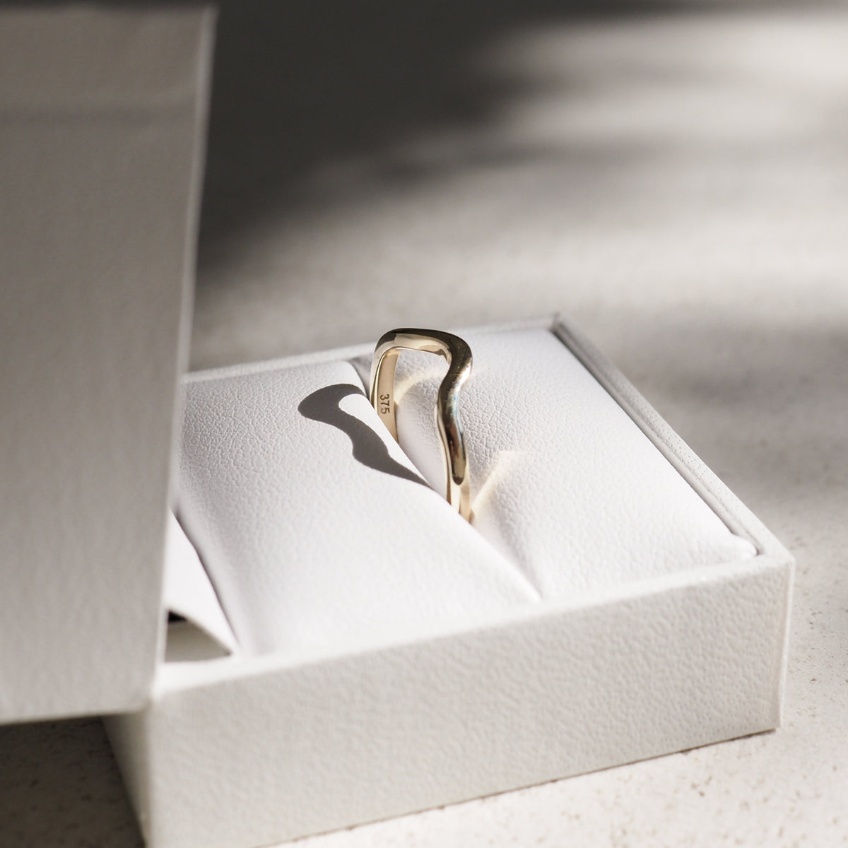Elegant gold ring with a wavy design resting in a white jewellery box, casting a soft shadow. Natural light enhances its luxurious appeal.
