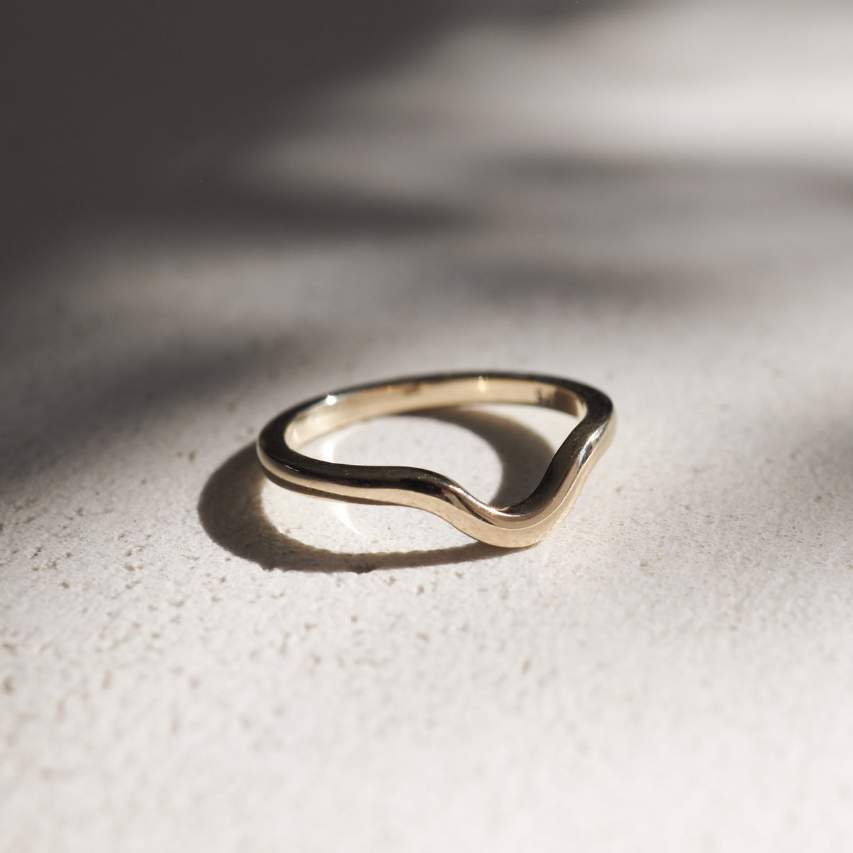 Gold wave ring on a textured surface, bathed in soft, natural light. Elegant curves reflect minimalist, luxury jewellery design.