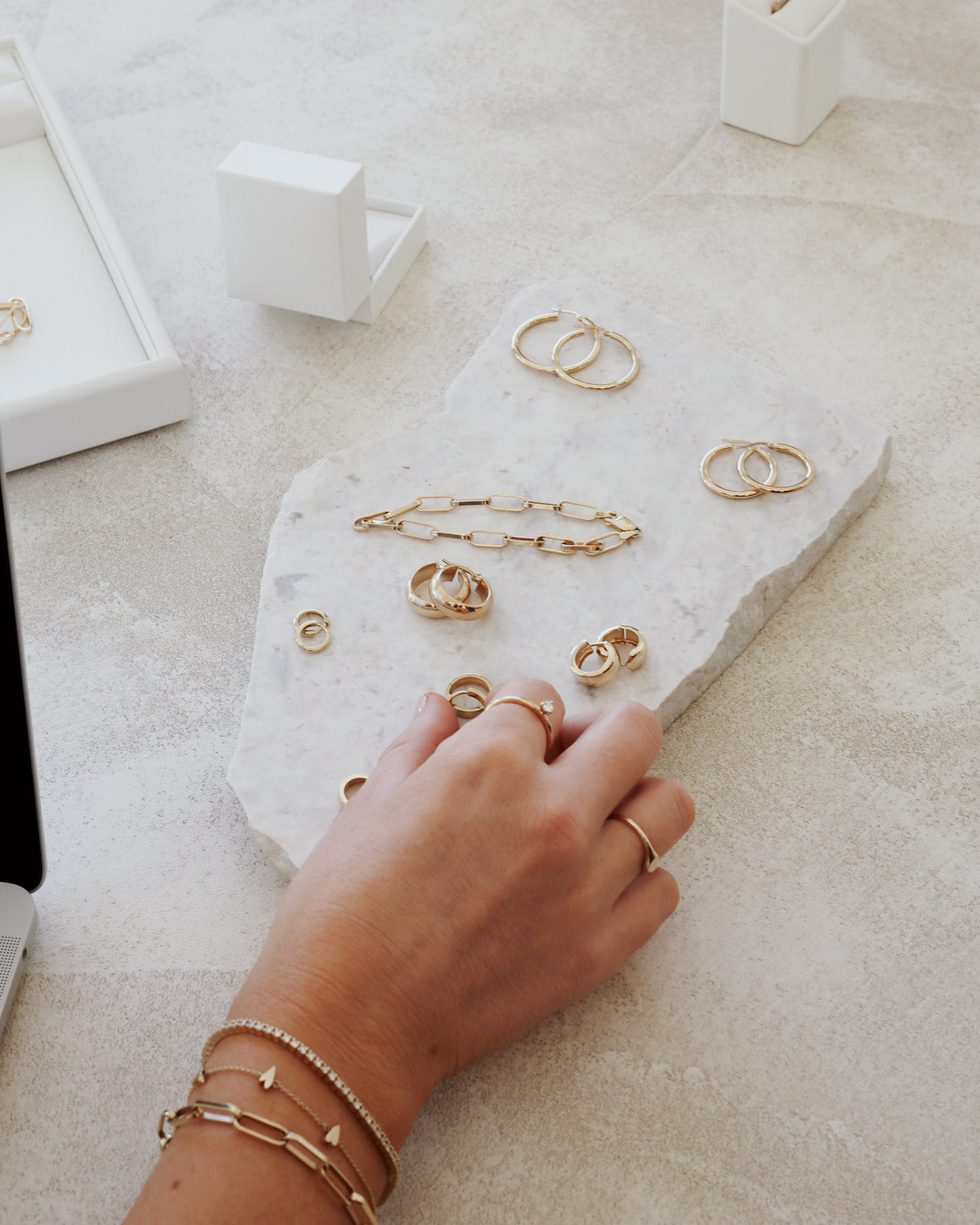 A hand adorned with gold rings and bracelets touches a marble slab displaying elegant gold jewellery, including rings and a chain bracelet.