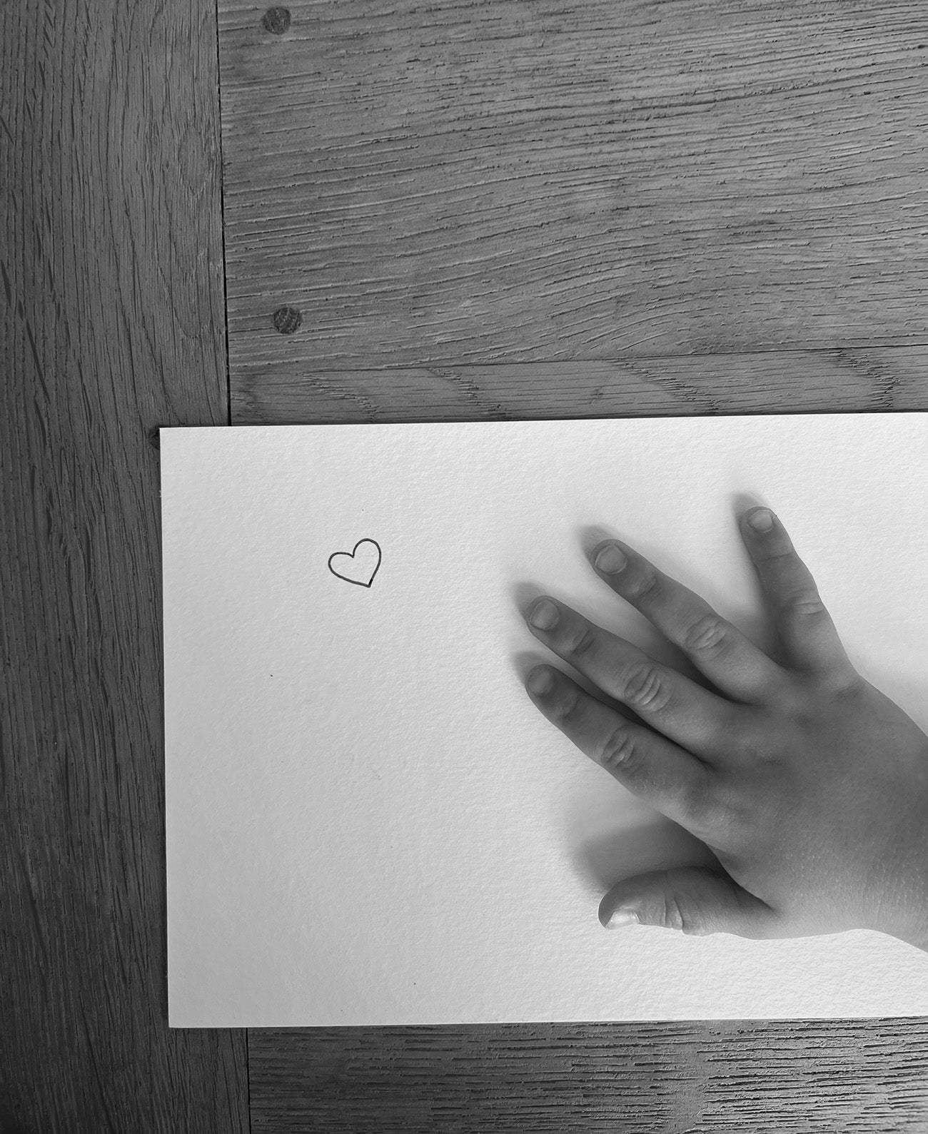 Child's hand on white paper with a small heart drawing, resting on a wooden table. Black and white image conveying simplicity and warmth.
