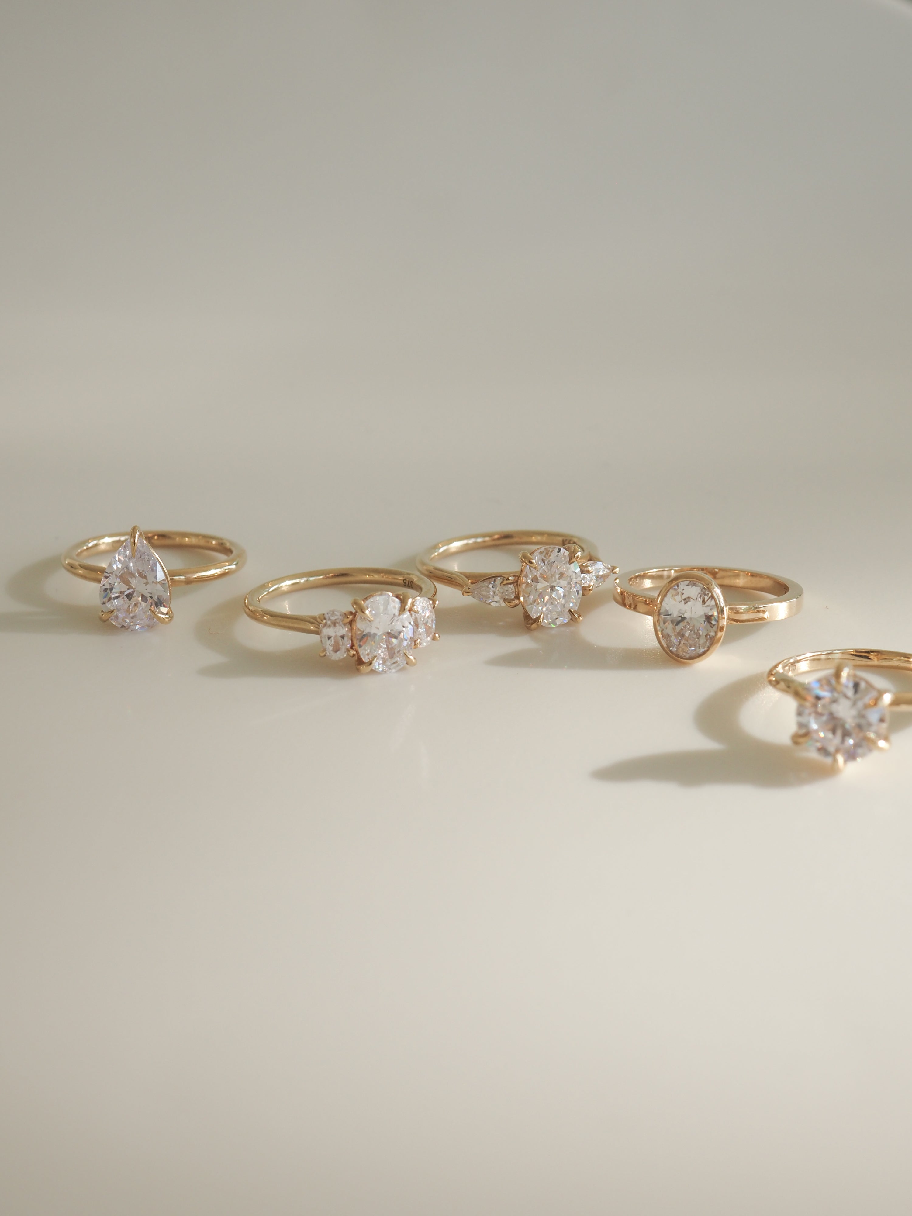 Six gold rings with various diamond cuts, including pear, round, and oval, elegantly displayed on a softly lit surface.